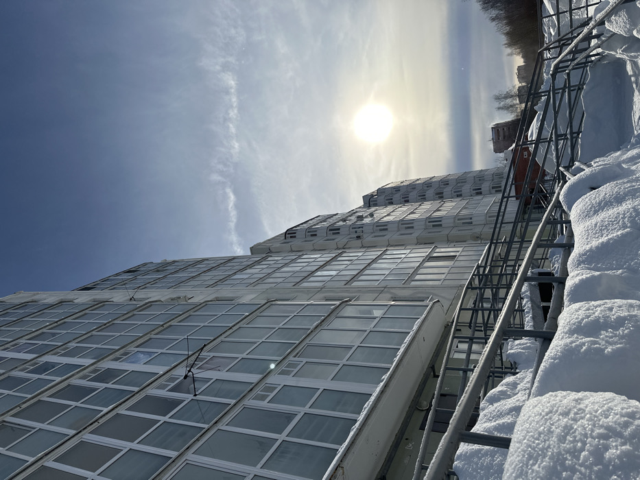 The image displays an urban residential environment in Perm, Russia, under clear winter conditions. A multi-story residential building, light in color, dominates the left and central portions of the frame, featuring numerous balconies enclosed with white-framed windows. Several antenna-like fixtures are visible extending from these balconies. Similar multi-story residential structures recede into the background on the right.
The lower half of the scene is covered in deep, undisturbed snow, forming significant drifts along the base of the foreground building and partially covering a set of dark metal railings, which delineate a cleared pathway or ramp. Bare deciduous trees are visible on the far right. The sky, occupying the upper portion of the image, is a light blue with minimal, wispy cloud formations. A prominent, un-obscured sun is situated in the upper-right quadrant, casting strong illumination across the scene and reflecting off the building's windows. No individuals or animate subjects are depicted.