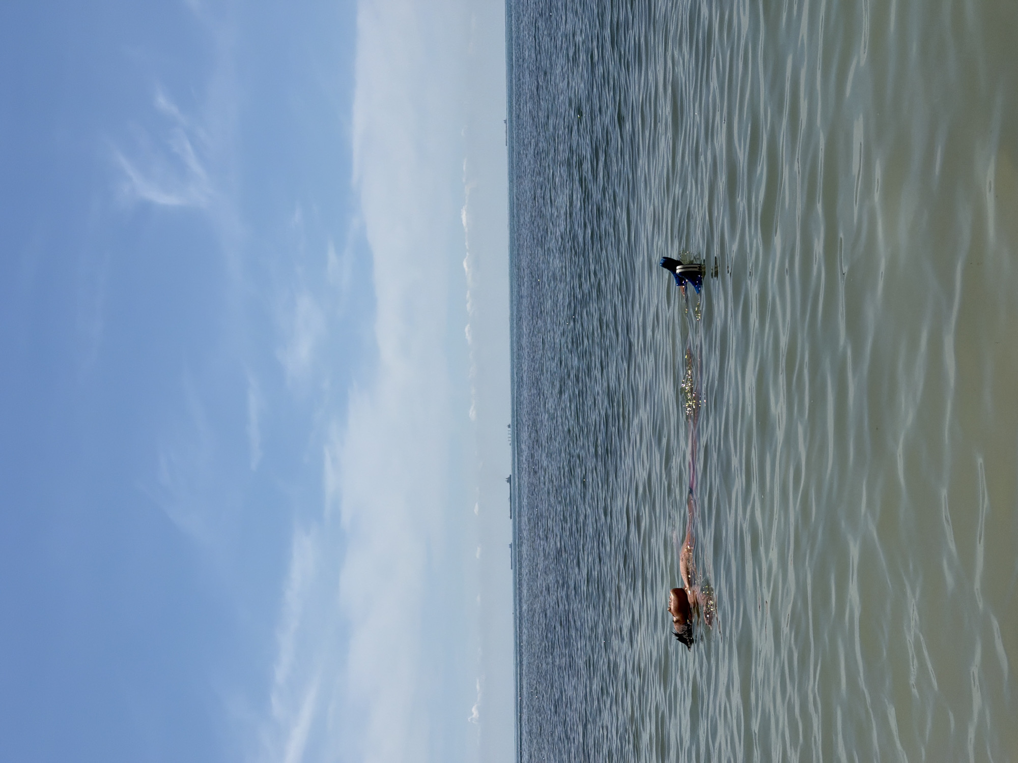 A person is floating on their back in the water, with their face tilted upwards towards the sky. They are wearing a snorkel mask on their face and flippers on their feet. The water is calm and the sky is blue with some scattered clouds. In the distance, several cargo ships are visible on the horizon. The foreground shows a shallow area of the water. The scene is outdoors and appears to be a warm, sunny day.