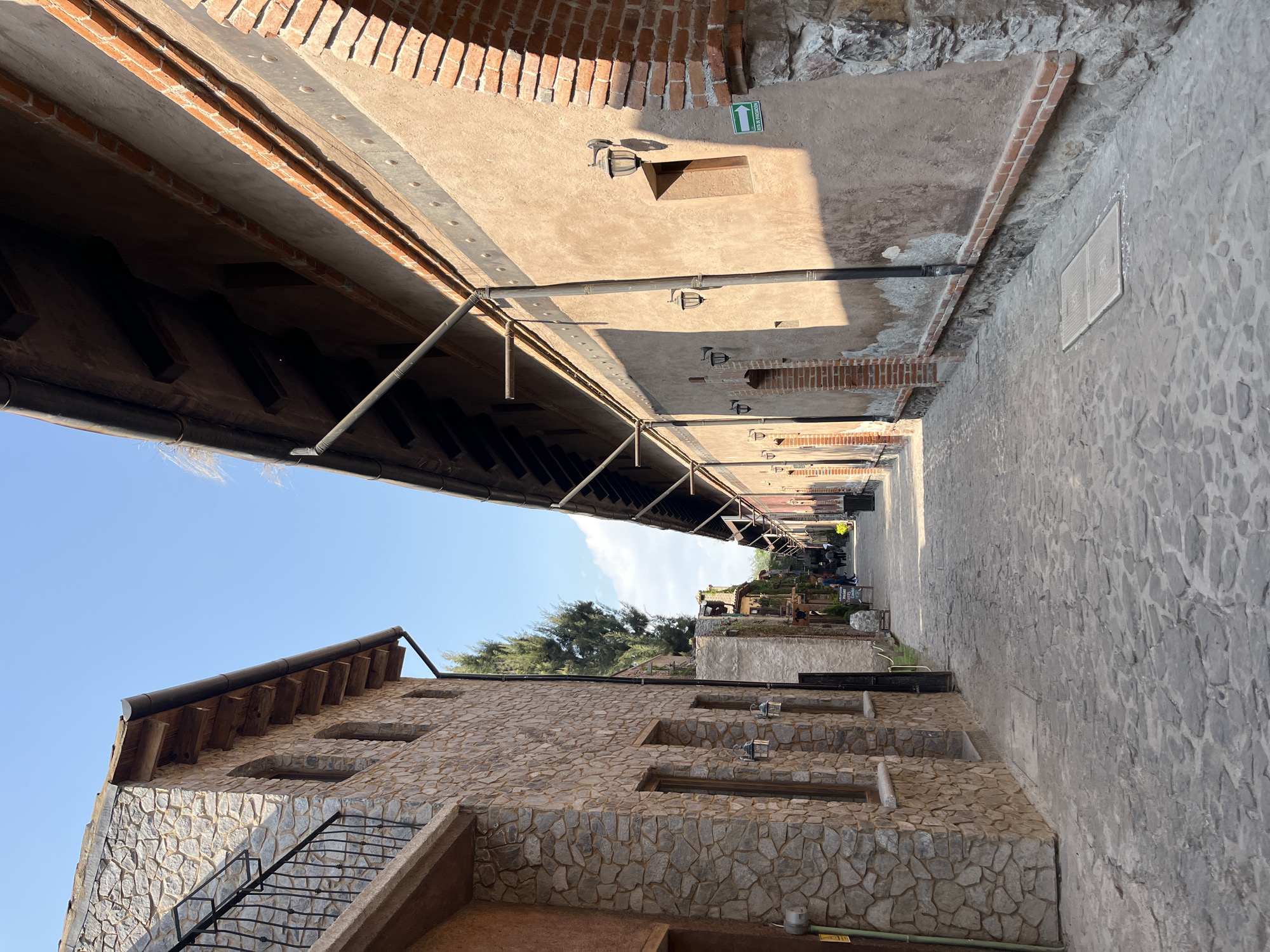 The image captures a narrow alleyway in Domingo Arenas, Mexico. The alley is flanked by buildings constructed with a combination of stone and brick. The building on the left features a stone facade and a staircase. A protective awning or structure covers the alley, supported by metal beams. In the distance, there are people walking and interacting in the alleyway, suggesting it is a public space. The environment is characterized by the architectural style, use of materials like stone and brick, and the presence of structures that offer shade.