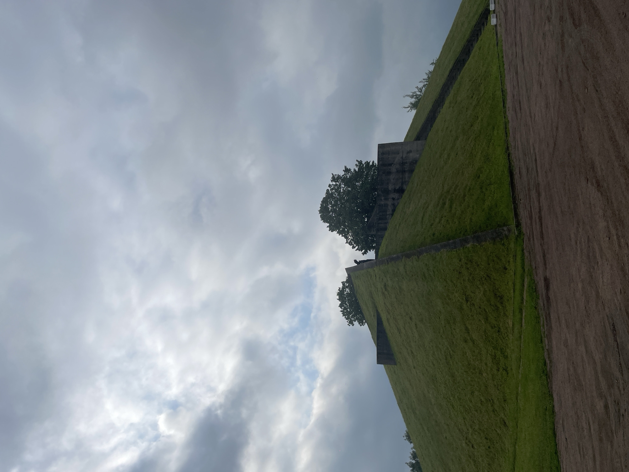 In Bol'shie Porogi, Russia, a large, grassy hill is topped by a structure with concrete walls and trees. The sky above is overcast with dramatic clouds. A single person stands near the top of the hill, facing away from the camera. The foreground consists of a dirt path.