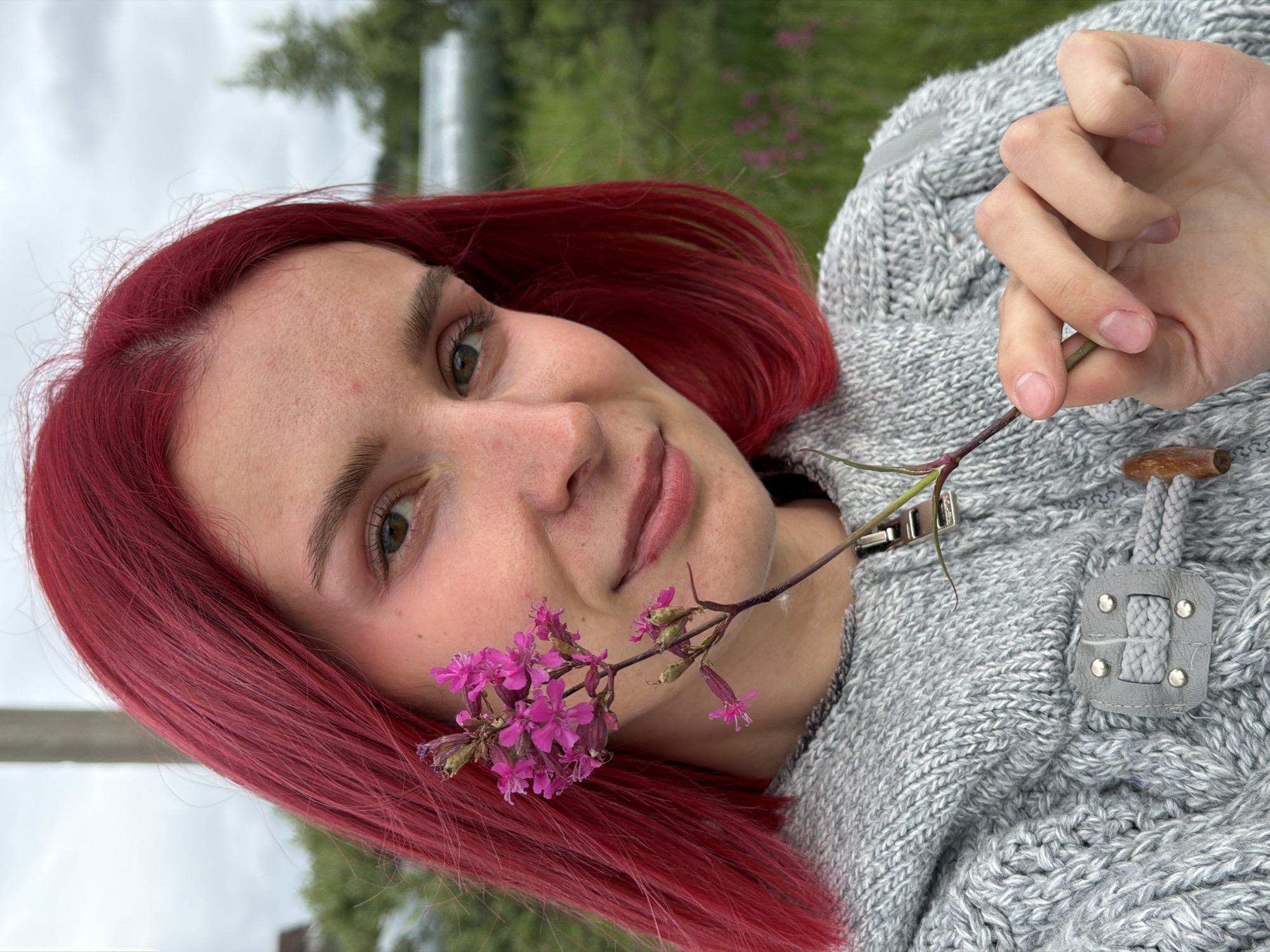 A young female with bright red, shoulder-length hair is depicted in a close-up portrait outdoors in the city of Chaikovsky, Russia. Her light-toned skin exhibits minor freckling across the nose and forehead. She has green-hazel eyes and light eyebrows, looking directly at the viewer with a slight smile. A subtle pink hue is visible on her eyelids and cheeks. She is wearing a grey, heavily textured knitted hooded sweater, possibly cable-knit, featuring a metallic zipper pull at the collar and a decorative grey fabric loop with a cylindrical wooden toggle closure on the upper chest. Her right hand, with natural unpolished fingernails, holds a single stem bearing multiple small, vibrant magenta-pink flowers and buds, positioned near the left side of her face. The background is blurred, showing a pale grey, overcast sky above and green foliage, trees, and indistinct light-colored structures in the mid-ground and distance. Faint pink flowers, similar to those held, are visible within the blurred green background on the right.