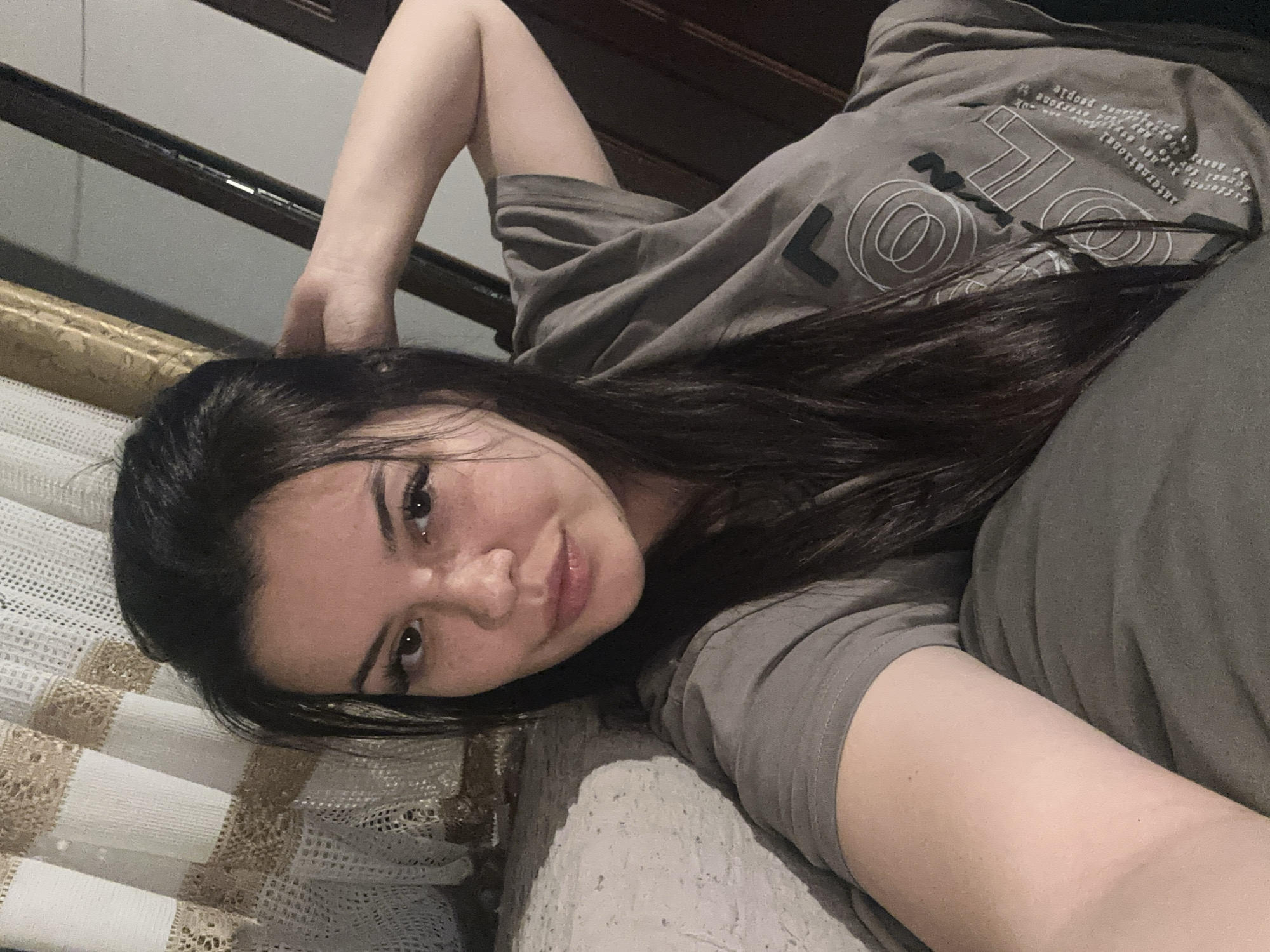 The image features a young person, likely female, with long, dark hair, lying down indoors. She is dressed in a short-sleeved, grey t-shirt and matching grey bottoms, suggesting casual wear for relaxing. Her head is resting on a textured, light-colored surface, possibly a bed or sofa, which is covered with a white and beige patterned blanket or sheet. The person is looking towards the camera with a gentle smile and a relaxed, content expression. Her left arm is bent, with her hand positioned near her head.
The grey t-shirt displays white graphic designs and text. Prominently visible are a large 'L' shape and a series of concentric circles. Smaller, partially obscured text can be deciphered, including words such as "everyone," "people," "new," "different," "heart," "international," "create," "explore," and "understand," arranged in an artistic, abstract pattern.
The background shows dark wooden furniture, possibly a bed frame or headboard, against a light-colored wall. The lighting appears soft and diffused, indicating an indoor setting, potentially during evening or night hours. There are no specific visual cues within the image itself to confirm the location as Chubra, Bulgaria. The central focus is on the person and her relaxed pose.