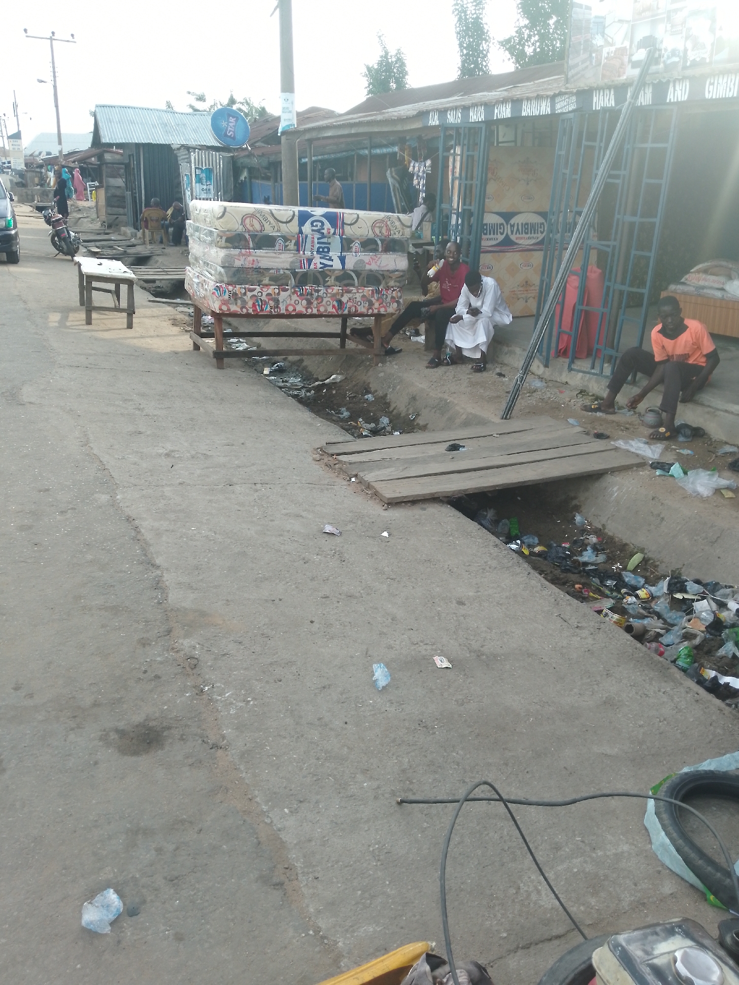 In Serti, Nigeria, a street scene unfolds with a focus on a mattress vendor's stall. A stack of mattresses, featuring colorful patterns and branding such as "GYIMBIYA GIMBA" and "BARUWA FOAM", is displayed on a wooden platform. Several individuals are present: a person in a bright orange shirt is seated on the ground near the stall, another in a white robe is sitting with a group of people, and two others are visible in the background. A motorbike is parked to the left, and a vehicle is partially visible. The environment shows a concrete pathway alongside a drainage ditch filled with debris, including plastic bottles and bags. A wooden plank serves as a makeshift bridge over the ditch. Signs with lettering like "HARA AM" and "GIMBIYA" are affixed to the stall's structure. The overall impression is that of a busy market area in Serti.