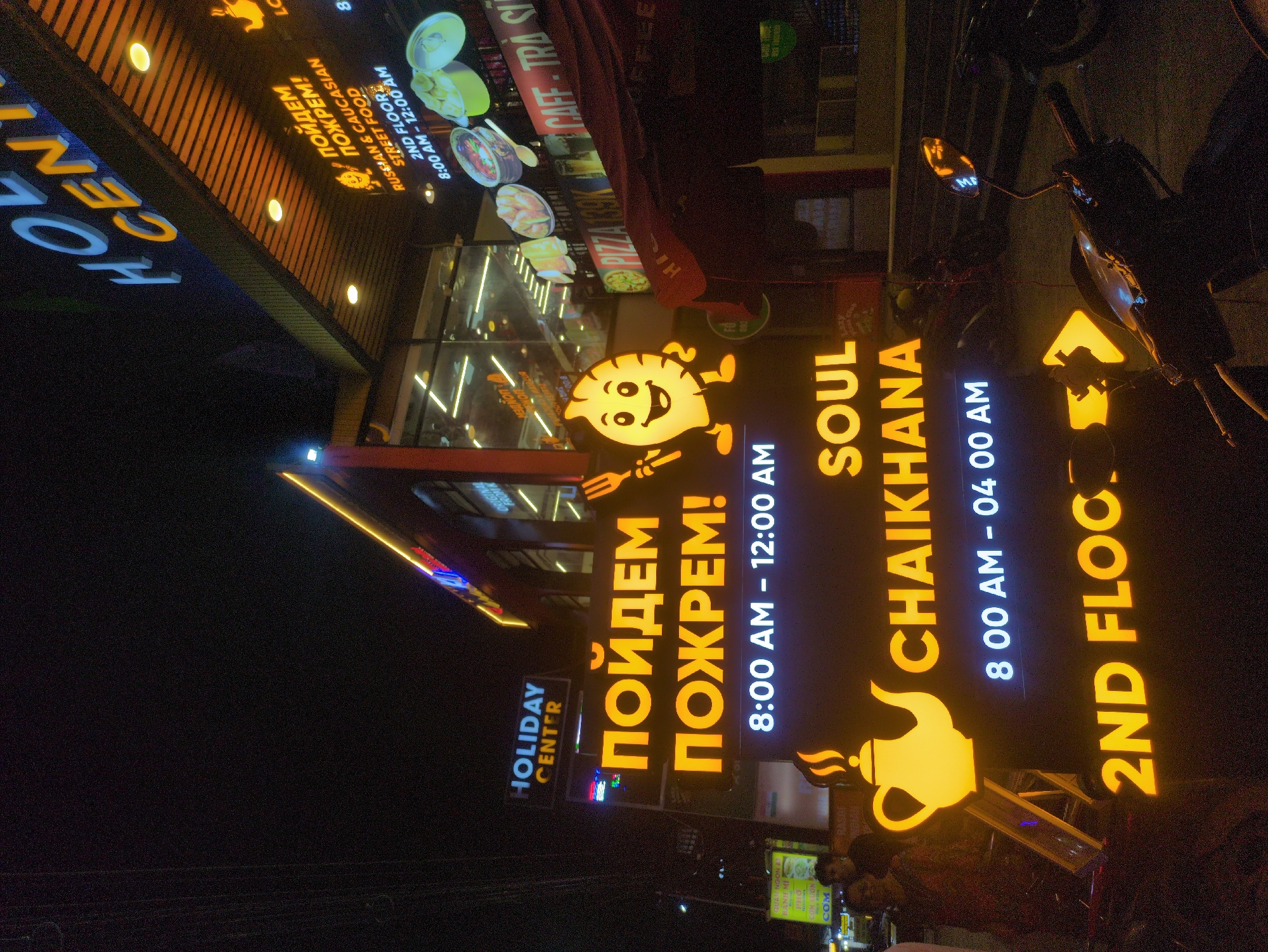 The image shows the exterior signage of "Soul Chaikhana" restaurant in Phú Quốc, Vietnam. There are two signs in Russian saying “Пойдем Пожрем!” or “Let’s go eat!” The signs indicate the restaurant is located on the 2nd floor of the Holiday Center, with the operating hours spanning from 8:00 AM to 4:00 AM. One sign says it serves Russian & Caucasian street food on the 2nd floor and is open from 8:00 AM to 12:00 AM. The signage incorporates playful imagery, including a smiling dumpling figure holding a fork and knife, as well as a teapot. A nearby scooter is parked and people are standing on the sidewalk in front of the restaurant. Other signs display the text “ORIGINAL Quan Ngon”, “Banh Mi Pho”, “Com Suon”, “COM” and “PIZZA 139K CAFE TRA SI”.