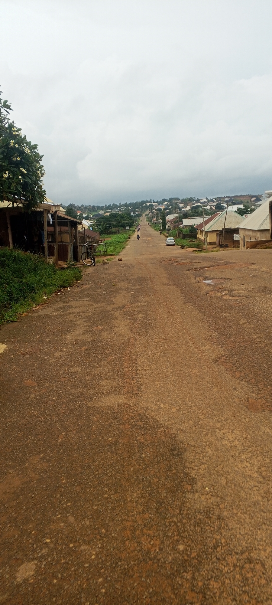 The image shows a long, sloping dirt road in Bahit, Nigeria. The road is lined with houses and vegetation on both sides. The sky is overcast, with a few clouds visible. On the left side of the road, there are some small structures with corrugated iron roofs and wooden walls. A bicycle is parked in front of one of these structures. Further up the road, there are a few people walking and a motorcycle. A white car is parked on the right side of the road. The houses appear to be made of mud bricks and have corrugated iron roofs. The environment is rural, with a sense of community and daily life.