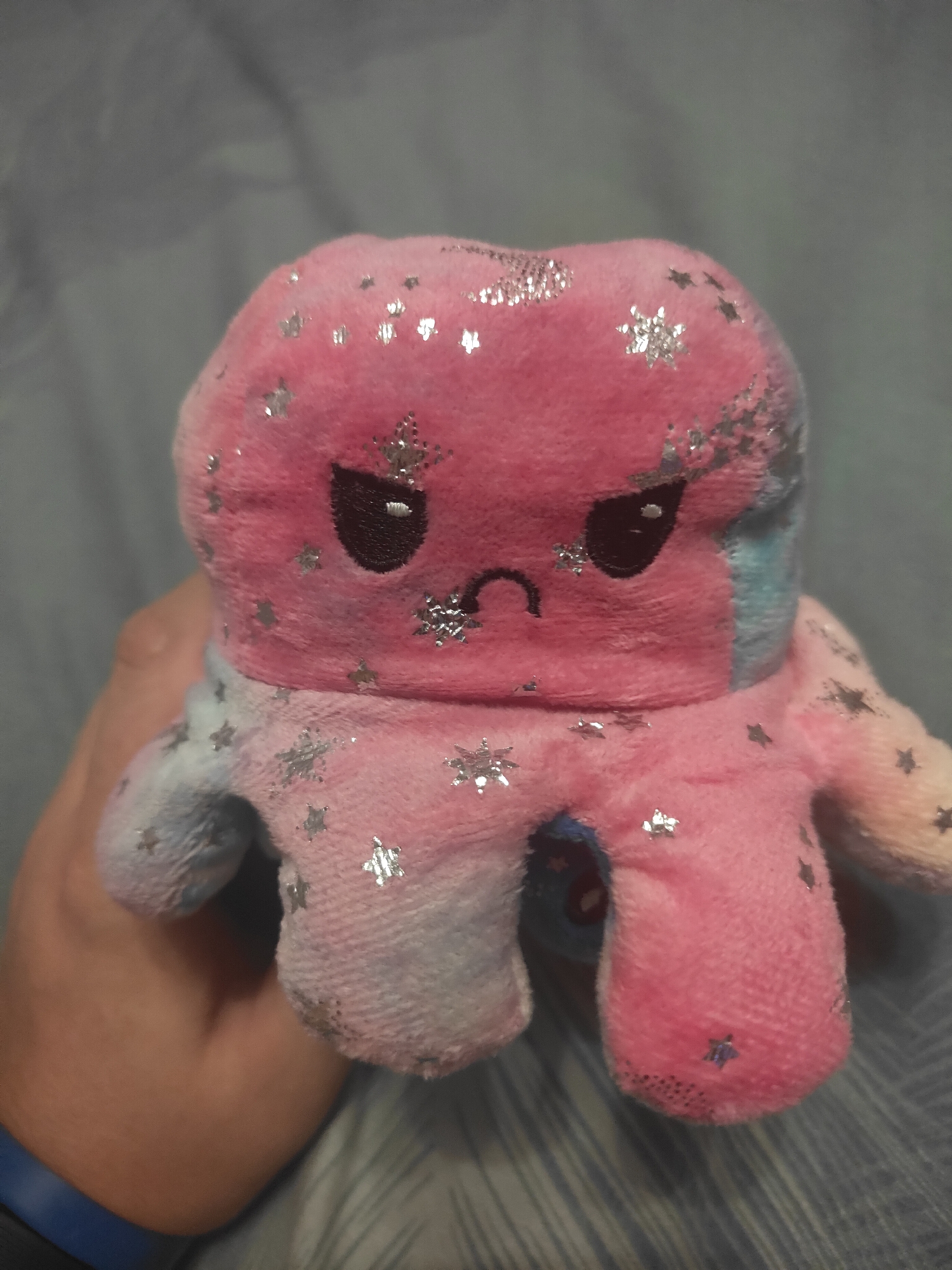 A close-up, medium shot shows a person's hand holding a pink and blue stuffed octopus toy. The octopus is plush and covered in small, silver star-shaped glitter accents. It has a round head with embroidered black eyes and a downturned mouth, giving it a grumpy or sad expression. The octopus's body transitions from pink on the top to blue and white on the bottom, with eight short tentacles extending downwards. The background is a softly blurred, greyish-blue, textured surface, possibly a blanket or bedspread. The lighting appears to be indoor, with no distinct shadows suggesting a specific time of day. The image is taken from a slightly high angle, looking down at the toy. No text is visible in the image.