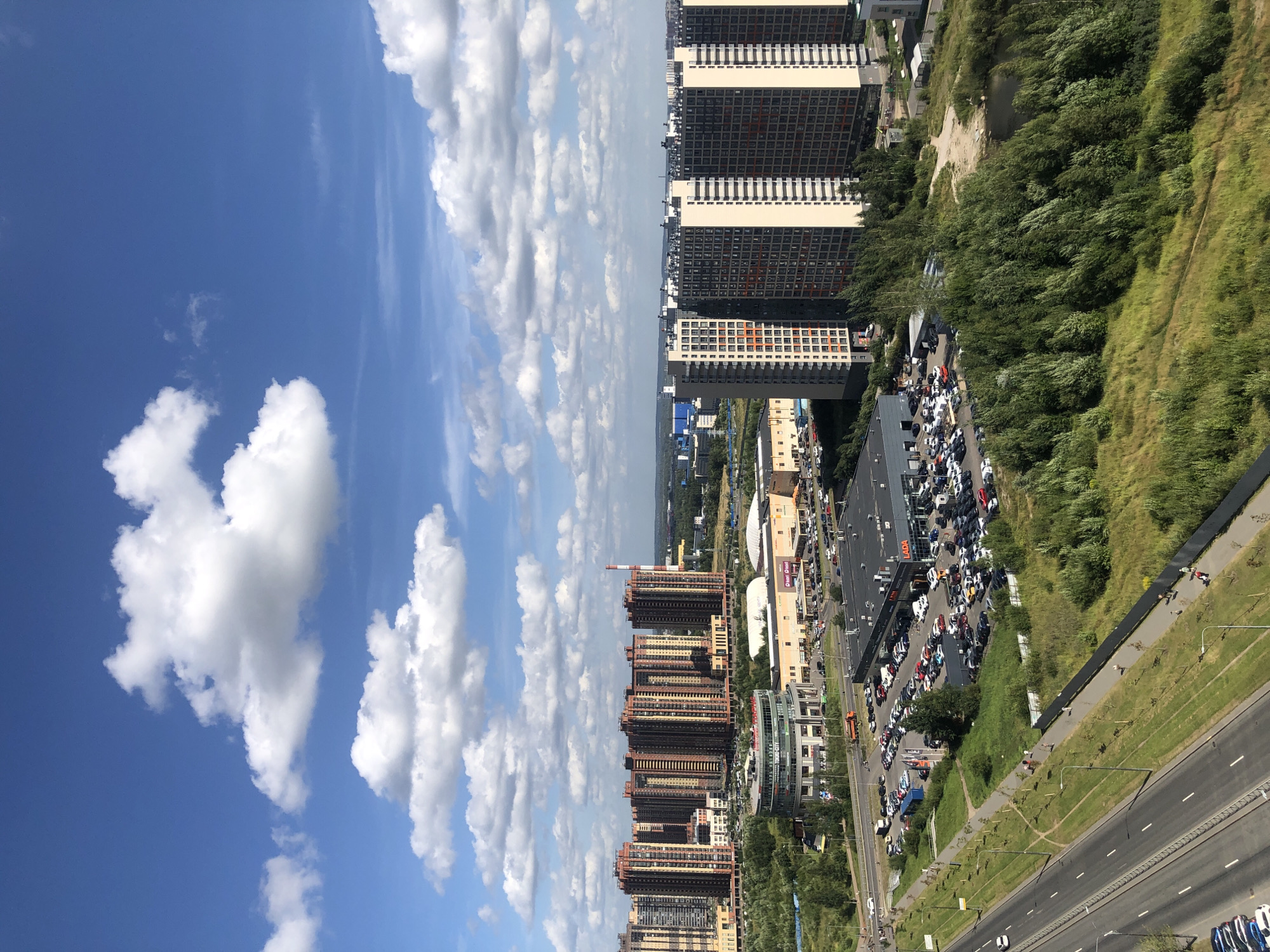 An aerial, eye-level view captures a sprawling urban landscape under a partly cloudy blue sky. The foreground features a paved road with a few cars. To the right of the road, a grassy embankment slopes upwards, transitioning into dense green trees. In the middle ground, a large, dark-colored commercial building, identified by a sign as "LADA," is surrounded by a nearly full parking lot filled with cars. Adjacent to this is a curved, multi-story building with a glass facade, and further back, several large, modern residential buildings with a reddish-brown and beige exterior dominate the skyline. In the distance, more buildings and a tall, red-and-white striped chimney are visible. The scene is bathed in bright daylight, suggesting it was taken on a clear summer day.