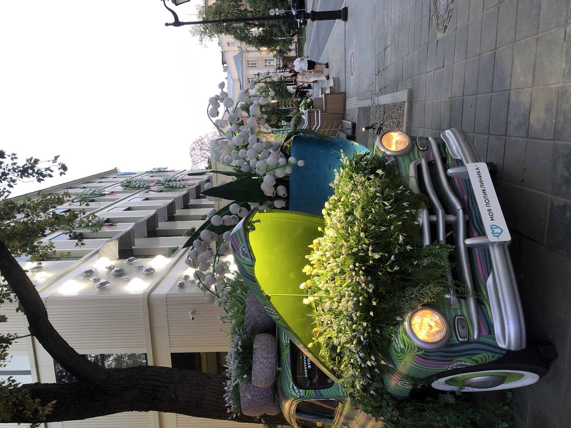 The image shows a vintage car decorated with flowers and plants on a sidewalk in Moscow, Russia. The car is green with colorful stripes and the hood is open, revealing a large bouquet of white flowers. The car is parked in front of a building with large windows and white vertical siding. A street lamp is visible on the right side of the image, and several people are walking on the sidewalk in the background. The overall scene appears to be a public art installation or decorative display.