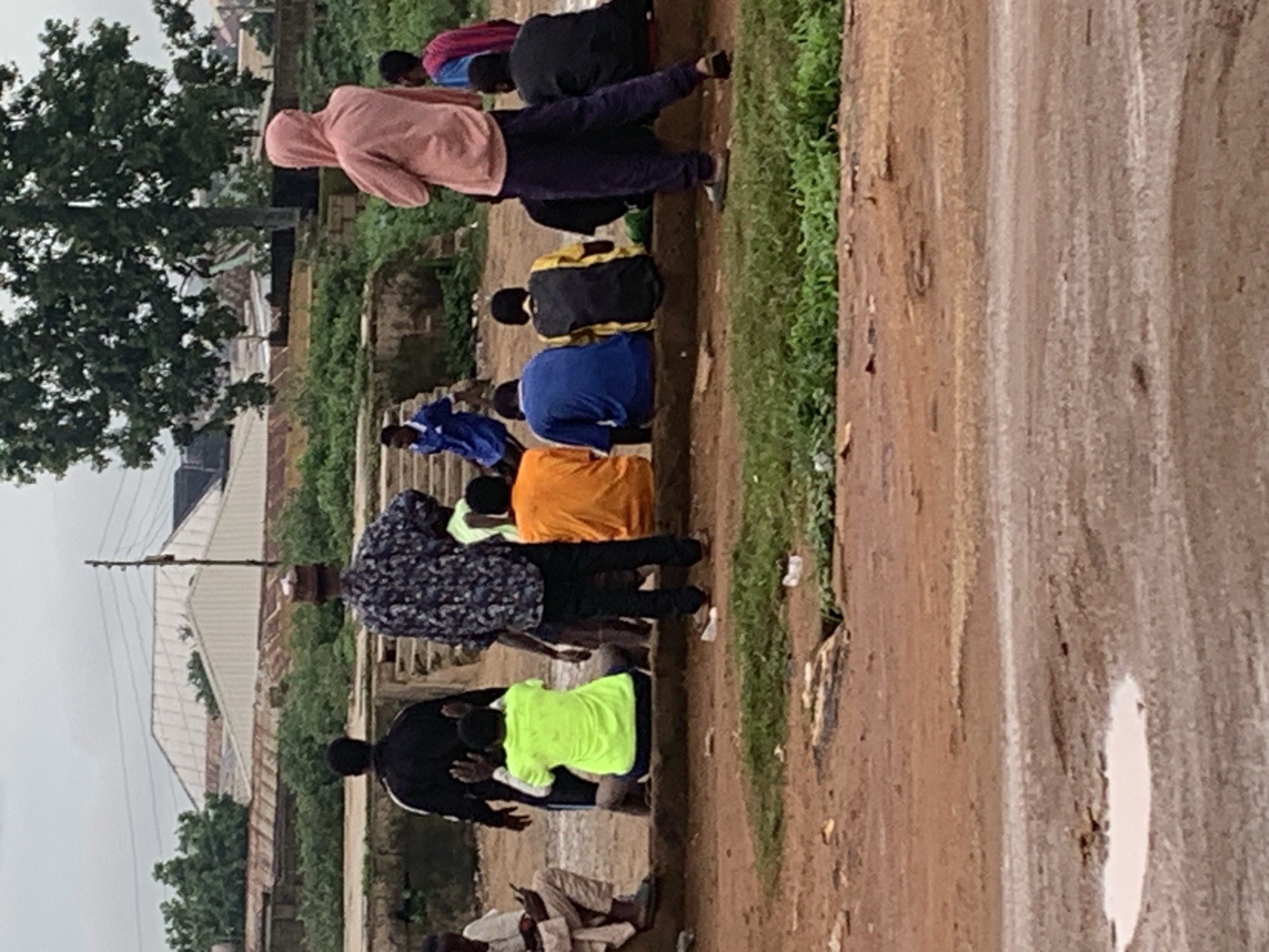 In Bukuru, Nigeria, a group of people are gathered on a raised dirt area. A man in a black jacket faces away from the camera, while another man in a patterned shirt and hat stands to his right. Several individuals are seated on a concrete ledge. One person wears a bright yellow shirt, another an orange shirt, a third a blue shirt, and a fourth a yellow and black shirt. A person in a pink hooded jacket stands on the far right, facing away from the camera. Another person wearing a blue shirt and shorts is in the background, and two individuals in dark clothing sit to the right of the seated group. The environment consists of dirt ground, grass, and some buildings in the distance.