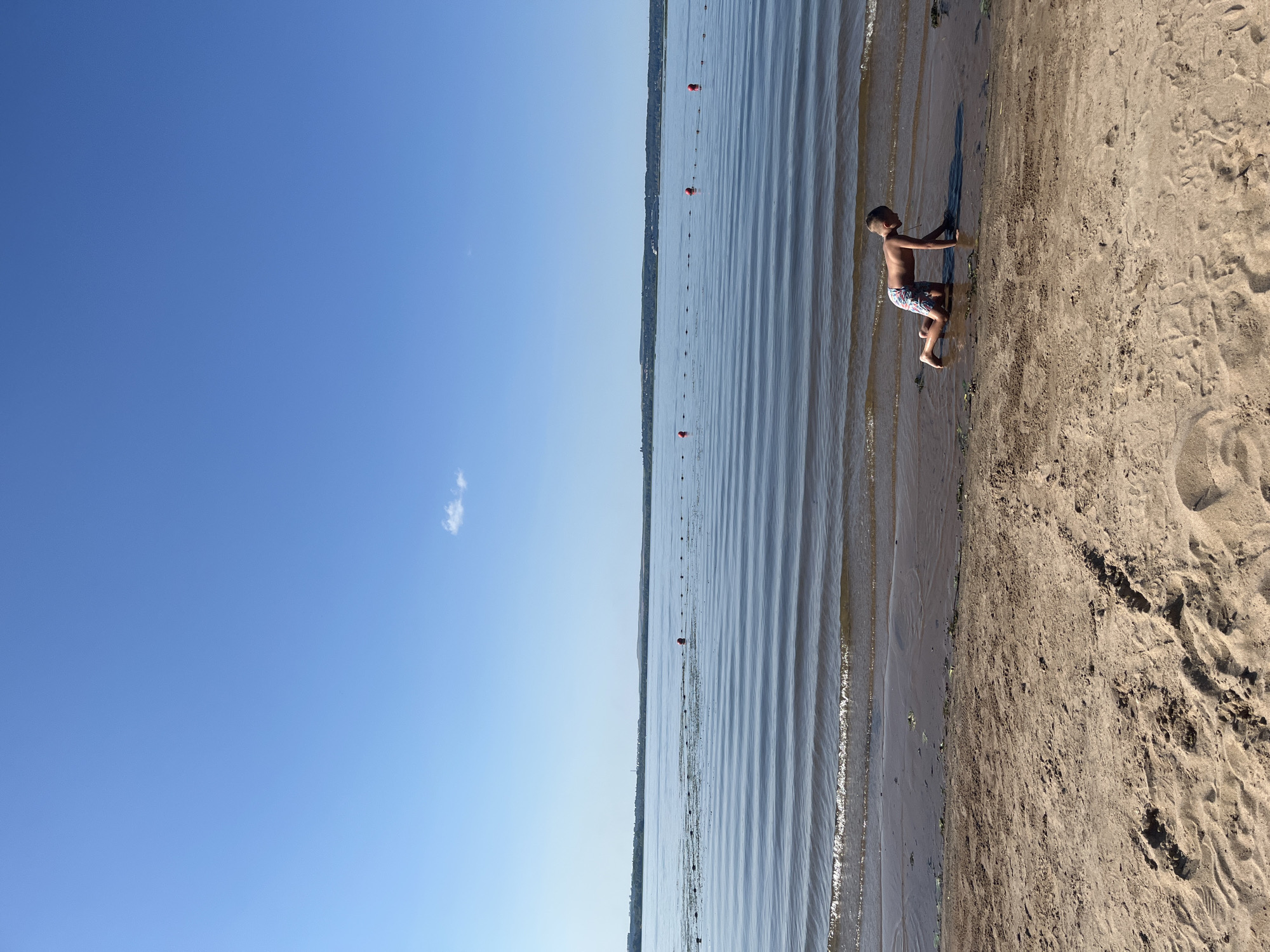 The image depicts a young boy on his hands and knees at the edge of a calm body of water, likely a lake or large river. The boy is wearing patterned swim trunks and appears to be interacting with the wet sand and shallow water. The foreground consists of a sandy beach with some debris and footprints. The water has gentle ripples and a line of red buoys stretches across it in the midground. The background shows a distant treeline and a hazy horizon under a clear blue sky with a single small cloud. The location is identified as the city of Shumeika, Russia.