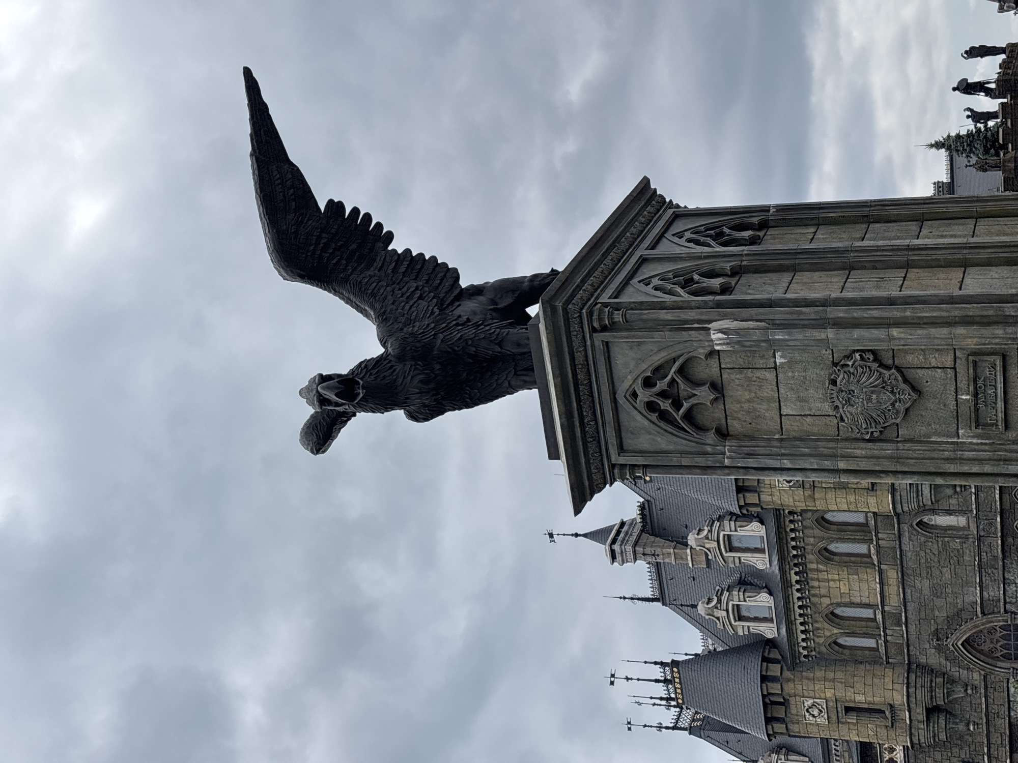 The image shows a large, dark-colored sculpture of a mythical creature, possibly a griffin, with outstretched wings and an open beak, perched atop a stone pedestal. The pedestal is ornate, with Gothic-style architectural elements and a coat of arms. In the background, a castle-like building with dark roofs, spires, and many windows is visible. The sky is overcast with gray clouds. The scene is located in Khryashchevka, Russia, at the "Castle of Garibaldi".