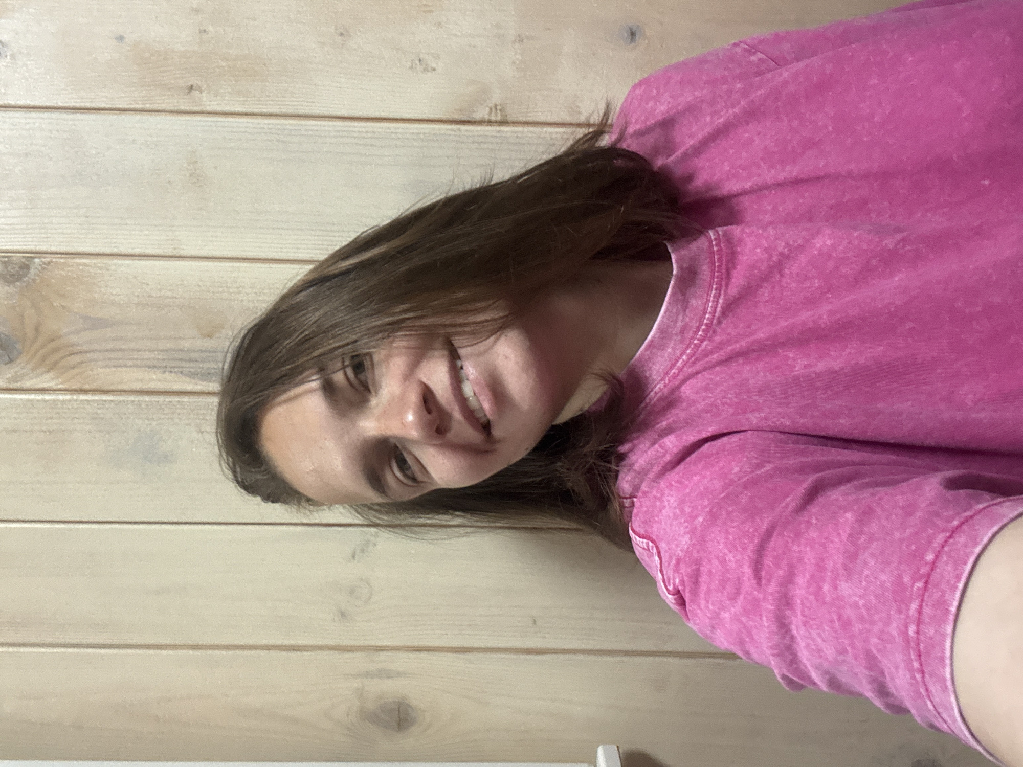 The image displays a young person, likely female, in a close-up portrait shot, taken indoors against a light-colored wooden wall. The subject has medium brown hair, which falls casually around her face, and is wearing a textured, faded pink t-shirt with a visible crew neck and seams. She is smiling broadly, with her lips slightly parted, and her eyes looking directly towards the camera, conveying a cheerful and relaxed emotion.
The background consists of horizontal wooden planks, suggesting a rustic or cabin-like interior. The lighting appears soft and even, indicating a daytime setting without harsh shadows. The person's head is slightly tilted, and the overall composition suggests a casual selfie or a candid moment. No specific activities other than posing for the photo are visible. There is no discernible text within the image. The location context is Раздолье, Russia.