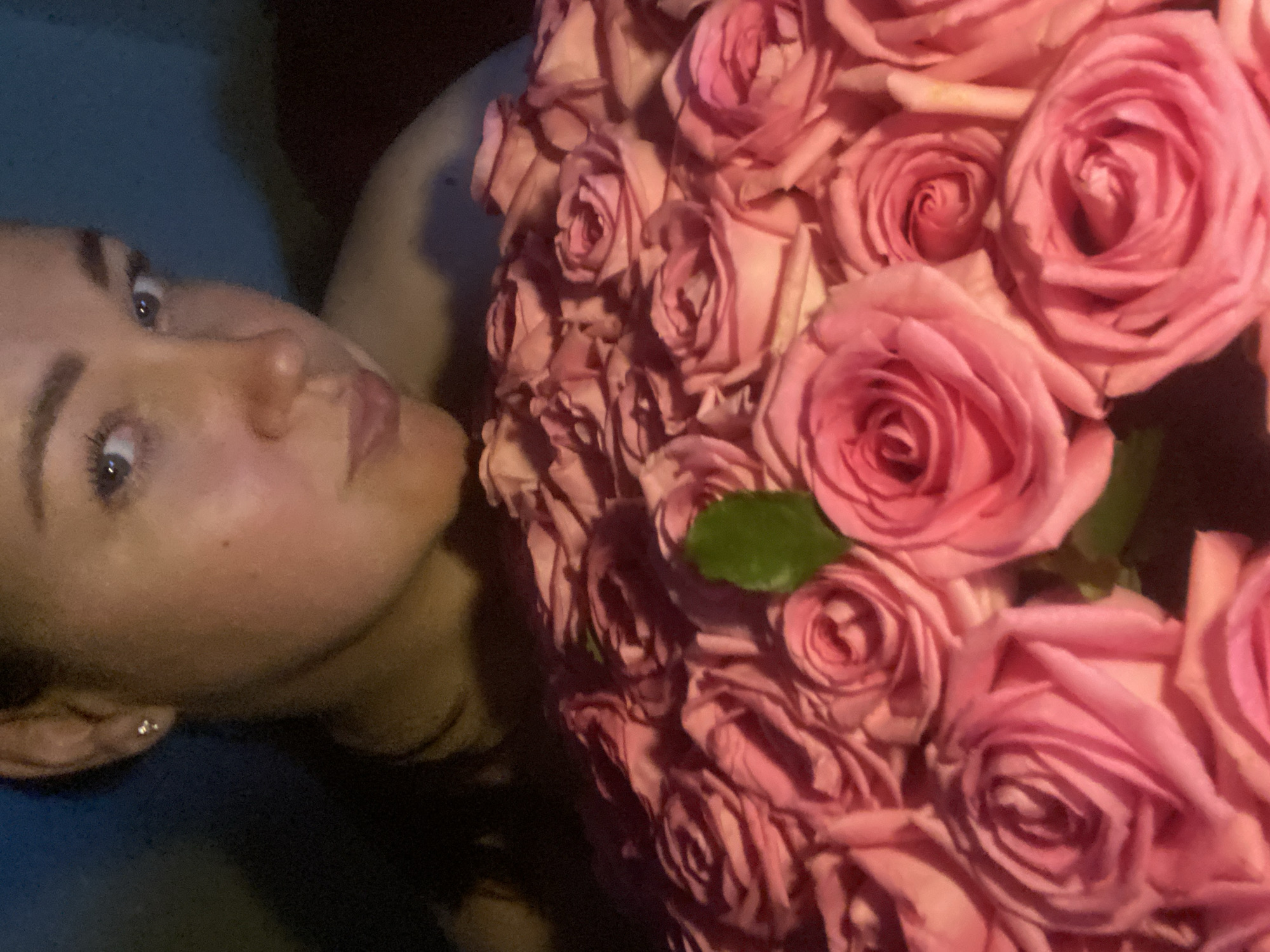 A woman with light skin, light eyes, and dark eyebrows is lying on her back, holding a large bouquet of pink roses. She is wearing an earring in her left ear. The background is dark and blurry, suggesting a low-light setting. The specific location within Sochi, Russia, cannot be determined from the image alone.