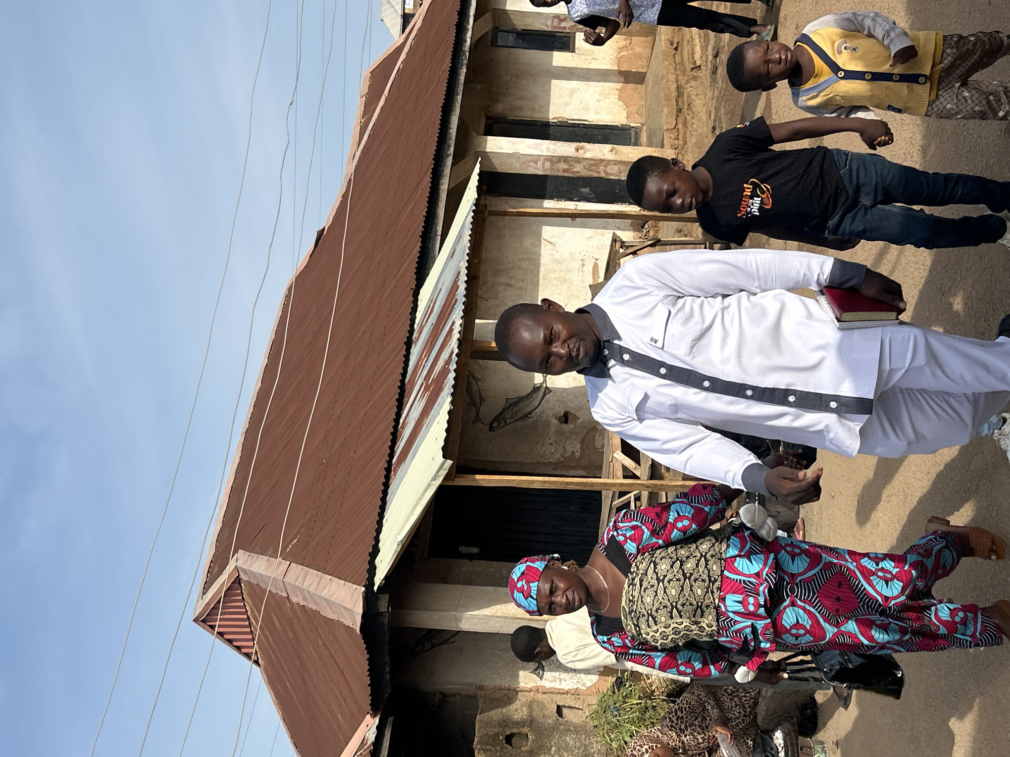 In Jos, Nigeria, a group of people walk along a dirt road in front of a row of buildings with corrugated metal roofs. A man in a white shirt and pants, holding a red book, walks in the foreground. To his left, a woman in a vibrant, patterned dress and headscarf carries a baby on her back. Two young boys, one in a black t-shirt and the other in a yellow cardigan, walk beside the man. The buildings appear to be simple structures, possibly homes or shops, with rough, textured walls. The sky is clear and blue, suggesting a sunny day. The overall scene depicts everyday life in an urban environment in Nigeria.