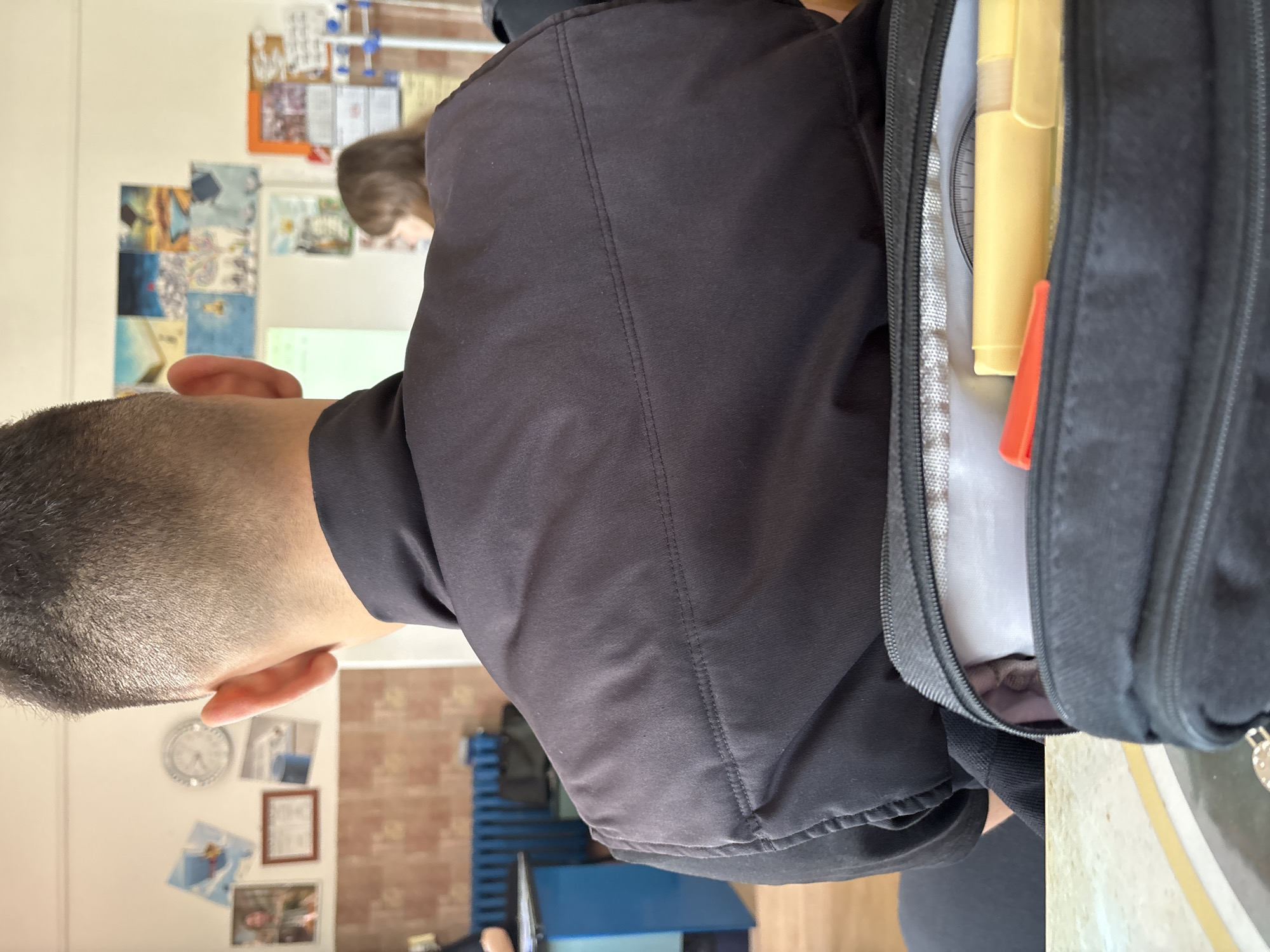 The image shows a person with short, cropped hair and light skin tone, likely in an indoor setting. The person is wearing a black jacket. Adjacent to the person is an open black backpack, revealing a protractor, a yellow highlighter, and an orange object inside. In the background are classroom-like elements, including posters and a clock on the wall. The scene appears to be a classroom setting, however, no definitive location within Panagyurishte, Bulgaria, is discernible.