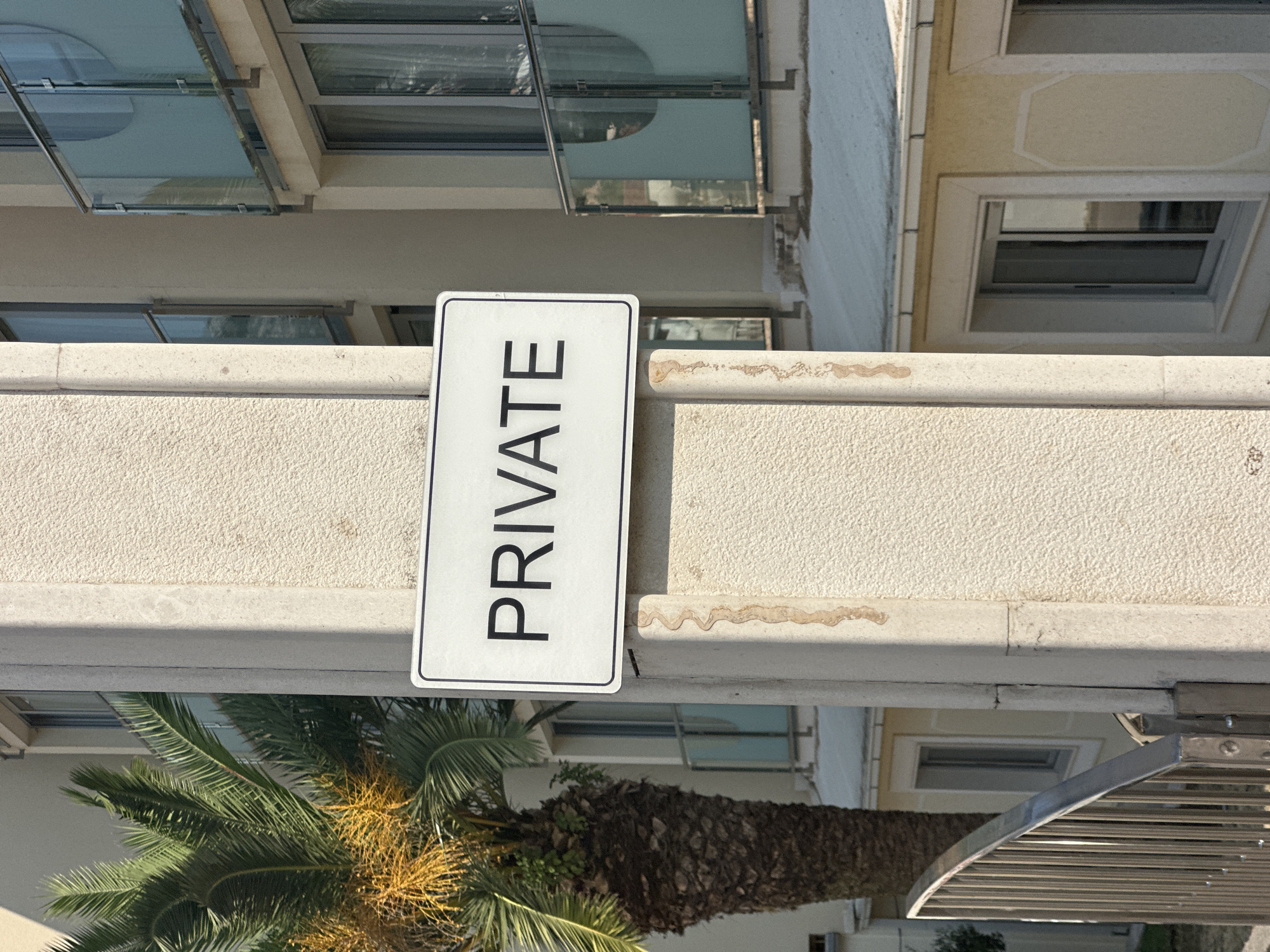 A sign with the word "PRIVATE" is attached to a concrete pillar in front of a building. The sign is white with black lettering. To the left of the pillar, a palm tree with yellow fruits is visible. The building behind the pillar has multiple windows and balconies with glass railings. The ground in front of the building appears to be covered in snow, which is unusual for the location, as Bečići, Montenegro, is on the Adriatic coast and typically experiences mild winters. The scene suggests a private property in an urban or resort area.