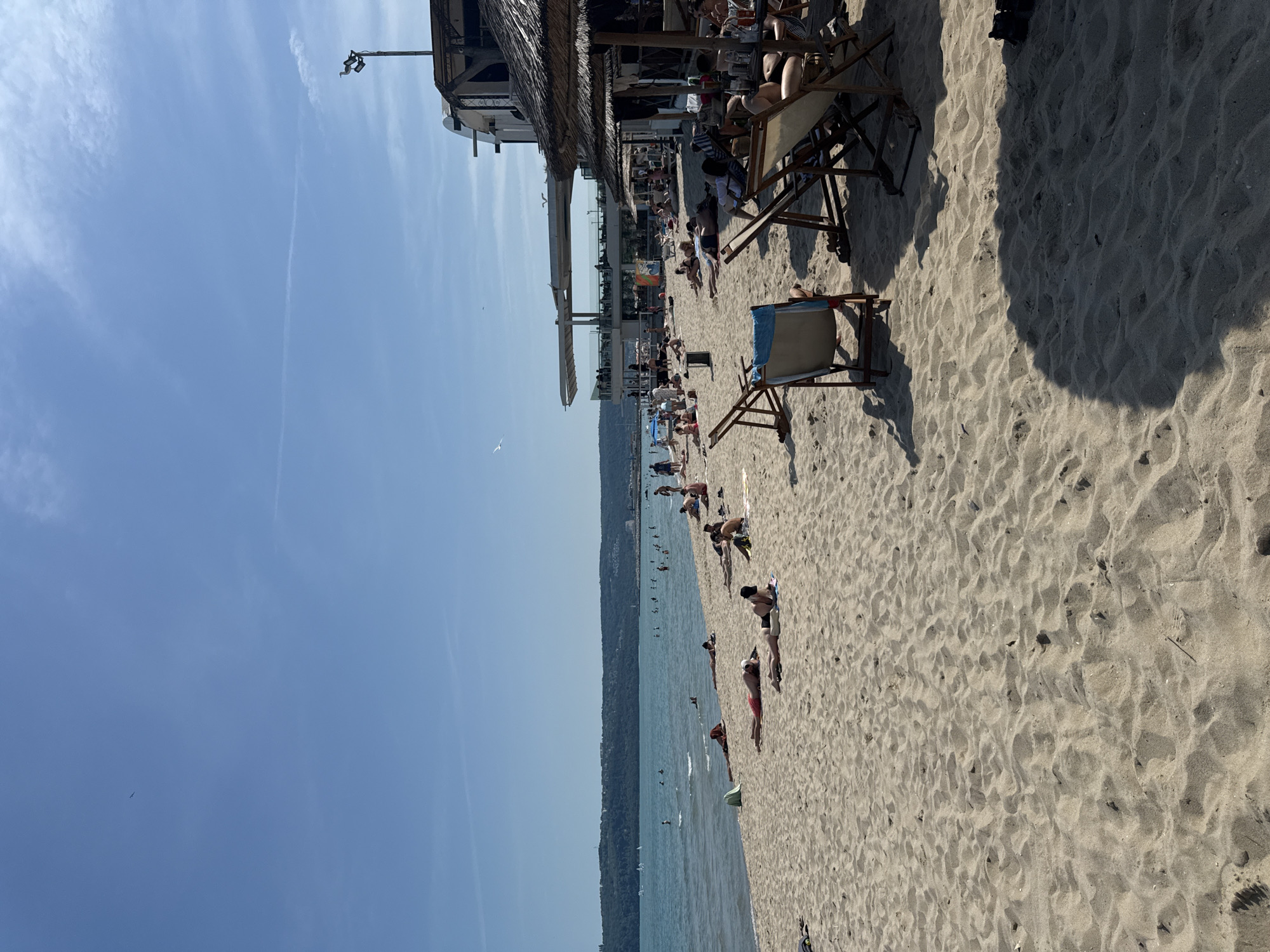 This full-shot image captures a sunny day on a sandy beach located in Varna, Bulgaria, specifically along the Varna Bay. The scene is dominated by a clear blue sky with minimal cloud cover and a faint contrail stretching horizontally.
In the foreground and midground, a light tan sandy beach is densely populated with individuals engaged in various activities. Numerous people are seen sunbathing, lying prone on towels directly on the sand, or reclining in wooden-framed deck chairs. Some beachgoers are partially obscured by the shadow of a large beach umbrella, while others are in direct sunlight. To the far right, a section of the beach operates as a shaded area with multiple occupied deck chairs and small tables, suggesting a beach bar or restaurant service, with drinks visible on tables.
The midground features the calm, blue-green waters of the Black Sea, with a gentle wave line near the shore. Many individuals are swimming, wading, or standing in the water. In the far distance on the water, small white sails indicative of windsurfing or small boats are visible. The distant shoreline across the bay shows a densely treed hill or mountain, punctuated by scattered buildings and urban development along its crest.
On the right side of the image, a prominent multi-level modern structure, likely a restaurant or club, with glass railings and a white facade, overlooks the beach. Its lower level appears to be a covered outdoor seating area with people. Adjacent to this structure, a traditional straw-thatched umbrella is visible, extending shade onto the beach. A light pole topped with what appears to be a seagull statue or a live bird stands near the building. Several live seagulls are seen flying in the sky, one distinctly visible near the modern building.
The overall atmosphere is one of leisure and activity on a warm, clear day at a popular beach destination.