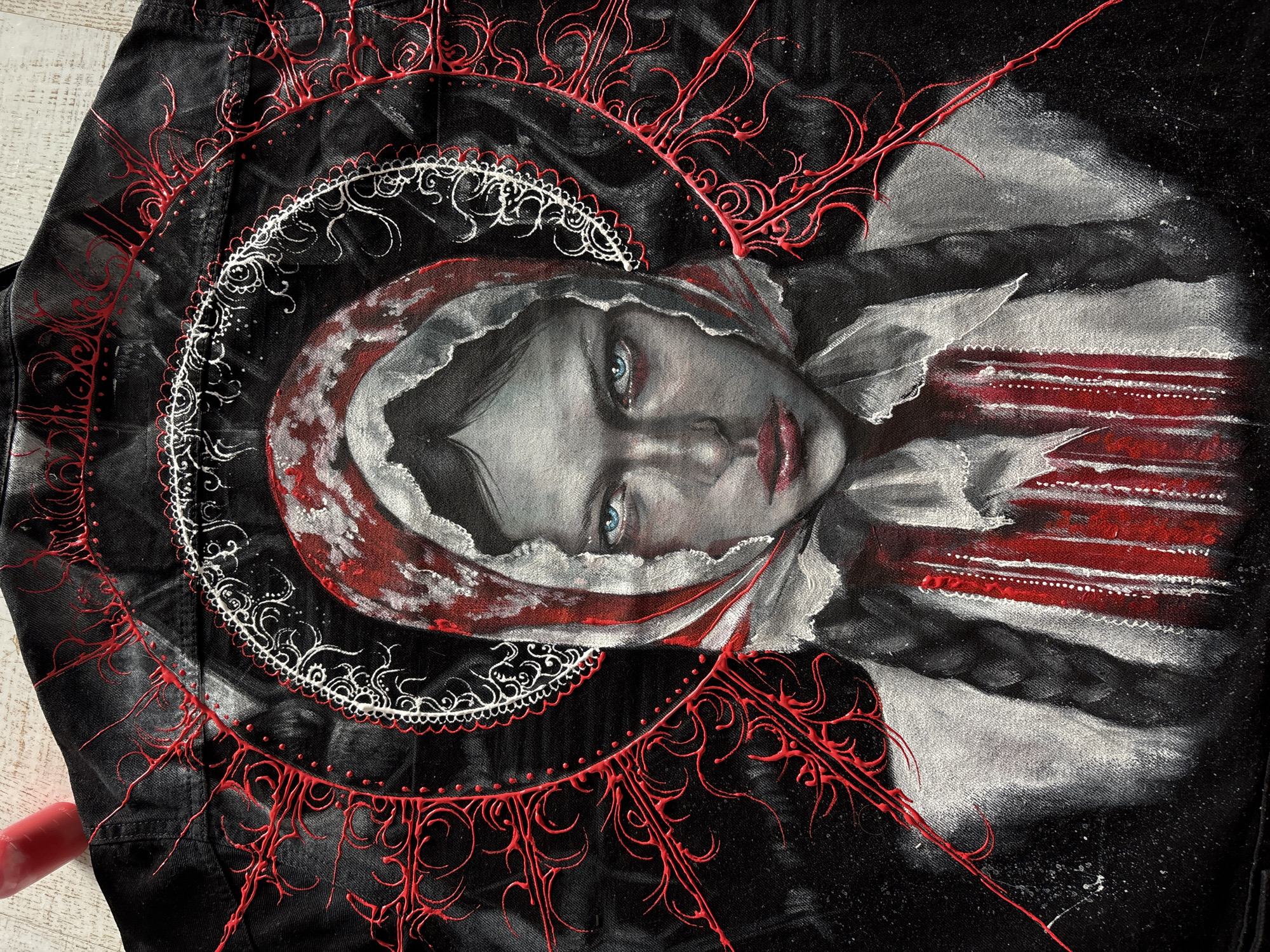 This media file displays a piece of wearable art, specifically a dark-colored garment, likely a black denim jacket, adorned with a detailed painting. The central subject is a portrait of a young woman with a solemn and direct gaze. Her bright blue eyes are a striking feature against her pale skin, and her lips are painted a deep red. She is depicted wearing a head covering, possibly a headscarf or veil, rendered in shades of red and white/grey.
Surrounding the portrait are ornate decorative elements. An inner circle of intricate white, lace-like patterns delineates the space around her head. Further out, a larger, organic-looking design in raised red lines branches outwards, resembling thorny vines or abstract tendrils, creating a textured contrast against the black fabric. On the right side of the portrait, bold, sweeping strokes of red and white/grey paint suggest a more abstract or dynamic background element, with the red areas having a somewhat stark, almost blood-like appearance.
The piece combines realistic portraiture with decorative and abstract elements, conveying an intense or melancholic mood. No activity is taking place; the jacket is laid flat, possibly on a light-colored wooden surface, suggesting it is being displayed or photographed. There is no visible text within the image. This media file was captured in Krasnoyarsk, Russia.