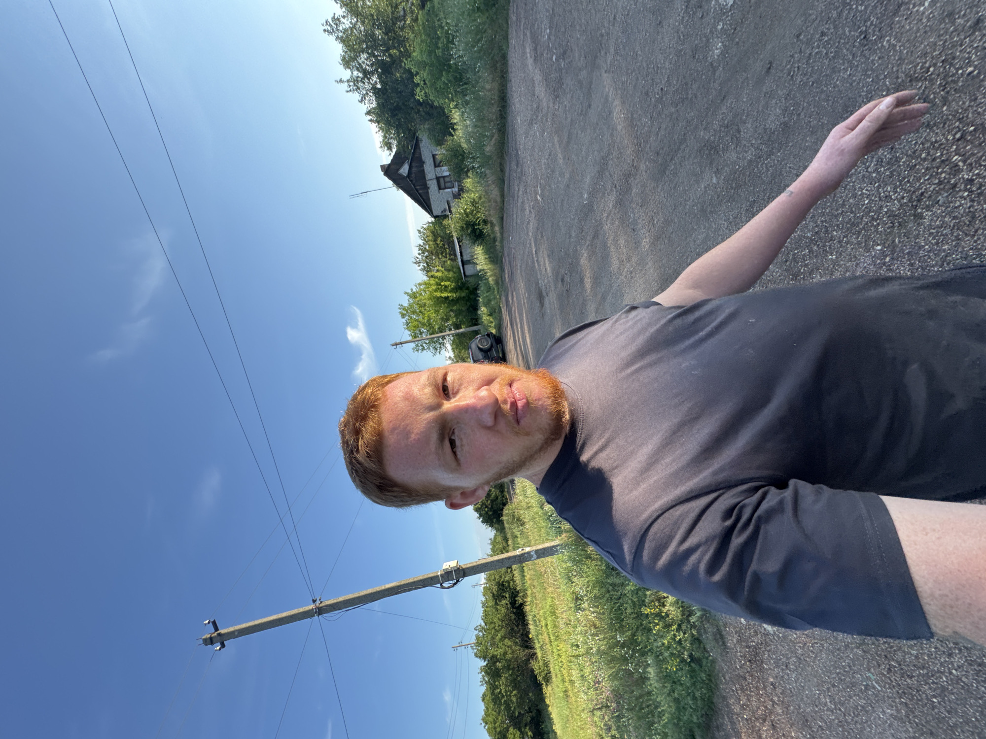 The image is a selfie taken outdoors in a rural or semi-rural area under a clear blue sky. A young man with red hair and a beard is in the foreground, wearing a dark t-shirt. He is standing on a gravelly dirt road. To his left, there is a grassy embankment with scattered wildflowers and a utility pole with electrical wires. In the background, there is a small, old house with a pitched roof, surrounded by trees and vegetation. A dark-colored SUV is parked on the road to the left of the house. The lighting suggests it is late afternoon or early evening.
