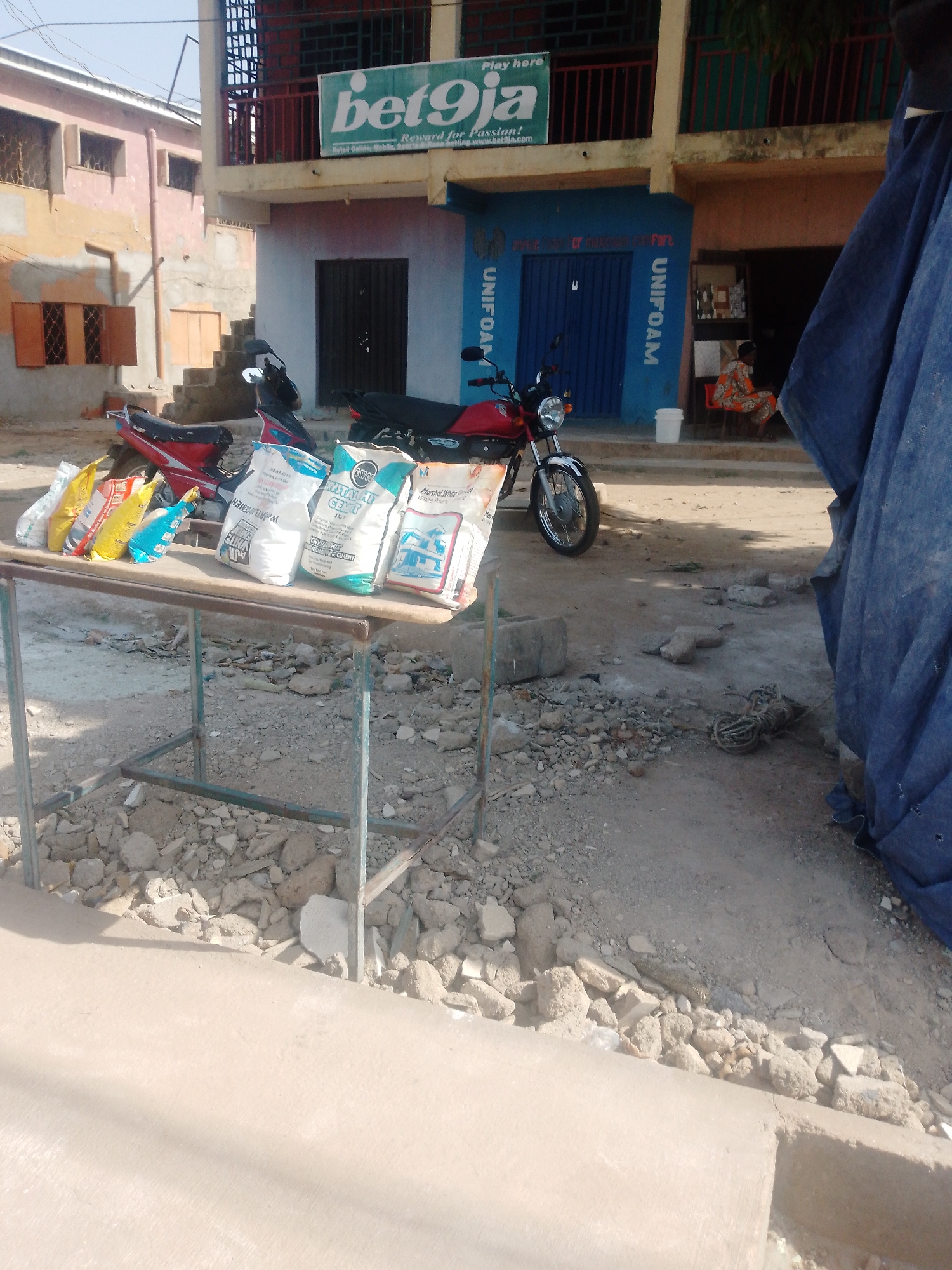 The image depicts an outdoor street scene in Sabon Tasha, Nigeria. A makeshift table displays bags of cement and other bagged goods for sale. Behind the table are two red motorcycles parked in front of buildings. One building has a "bet9ja" sign indicating a betting shop. Another building is labeled "UNIFOAM". A person is sitting inside an open doorway, visible behind the motorcycles. A blue tarp is partially visible on the right side of the image. The street surface is rough, covered with loose debris and gravel.