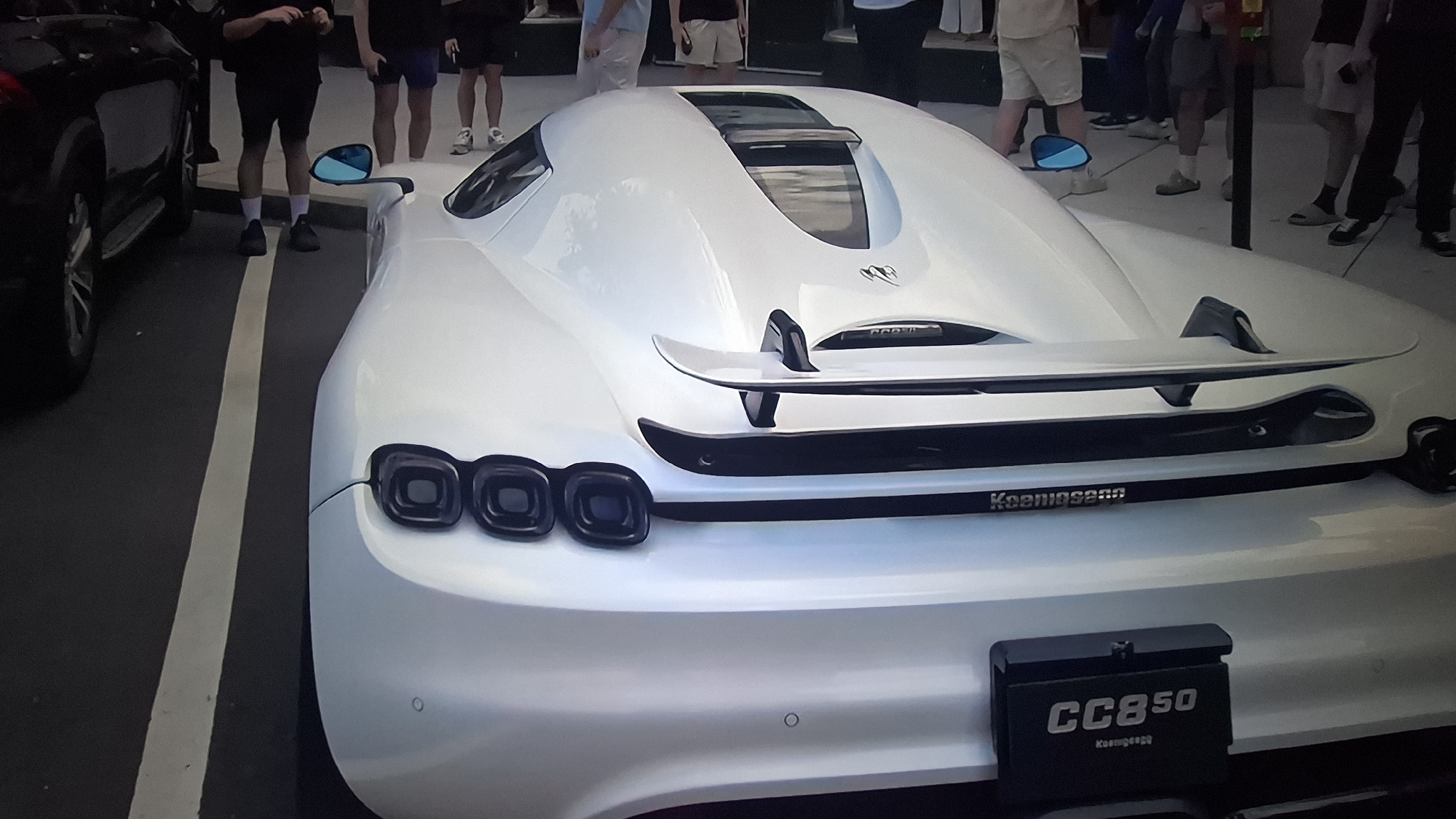 The image captures the rear view of a pristine white Koenigsegg CC850 hypercar, parked outdoors on an asphalt surface. The car dominates the frame, showcasing its distinctive design elements. Notable features include triple circular taillights on each side, a prominent rear spoiler, and blue-tinted side mirrors. The engine cover has a sleek, aerodynamic shape with a central transparent panel and the stylized Koenigsegg logo.
Several people are visible in the background and around the car, mostly showing their legs and lower torsos. They appear to be standing and observing the vehicle, suggesting a car show, exhibition, or public display event during the daytime. On the far left, the front wheel and part of a black SUV or car are visible, parked parallel to the Koenigsegg. White painted lines on the asphalt separate the parking spaces.
Visible text includes "Koenigsegg" spelled out on the lower rear bumper, and "CC850" prominently displayed on the black license plate holder, with "Koenigsegg" in smaller script underneath it. The lighting is bright and even, indicative of clear weather conditions. The overall scene conveys an impression of admiration for the high-performance automobile.