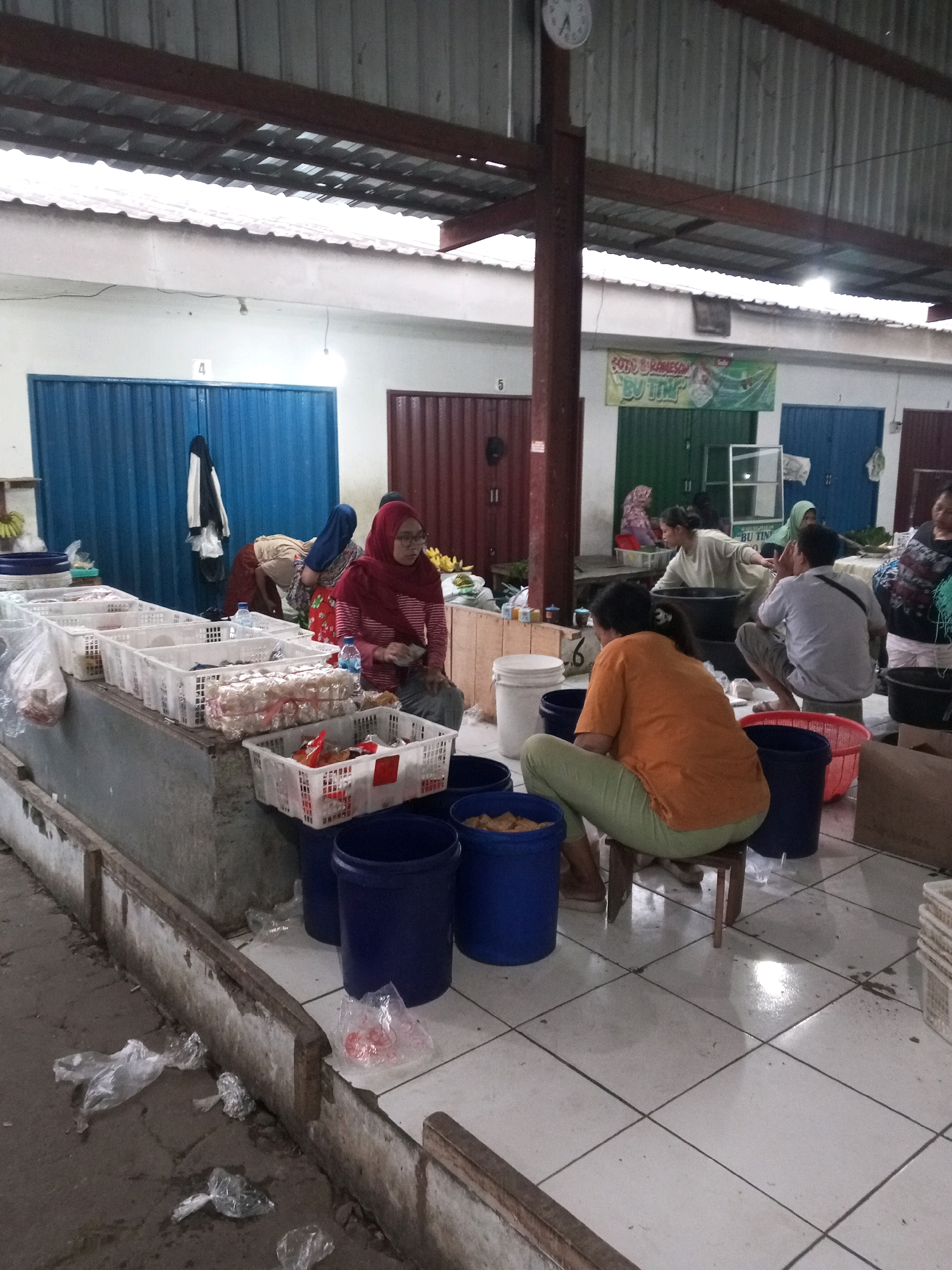 This image depicts a bustling marketplace in Gentan, Indonesia. In the foreground, a woman in an orange t-shirt and green leggings is seated on a small wooden stool, facing away from the camera. She appears to be engaged in an activity involving a large blue bucket. To her left, another blue bucket is visible, containing what looks like food items. Behind her, several other individuals are present, including a woman in a red hijab and glasses who is sitting at a stall laden with white plastic baskets filled with various goods, possibly food products. Other people are seen standing and interacting in the background, suggesting a dynamic market environment. The marketplace itself is covered by a corrugated metal roof supported by metal beams, with white walls visible in the background. Blue and red metal doors line the stalls. The floor is tiled with a white and gray checkered pattern, with some areas appearing wet. Plastic bags, some containing items, are scattered on the ground, indicating typical market activity and potential cleanup needs. A clock is visible on the wall near the ceiling. The overall scene conveys a sense of local commerce and daily life in a lively Indonesian market.