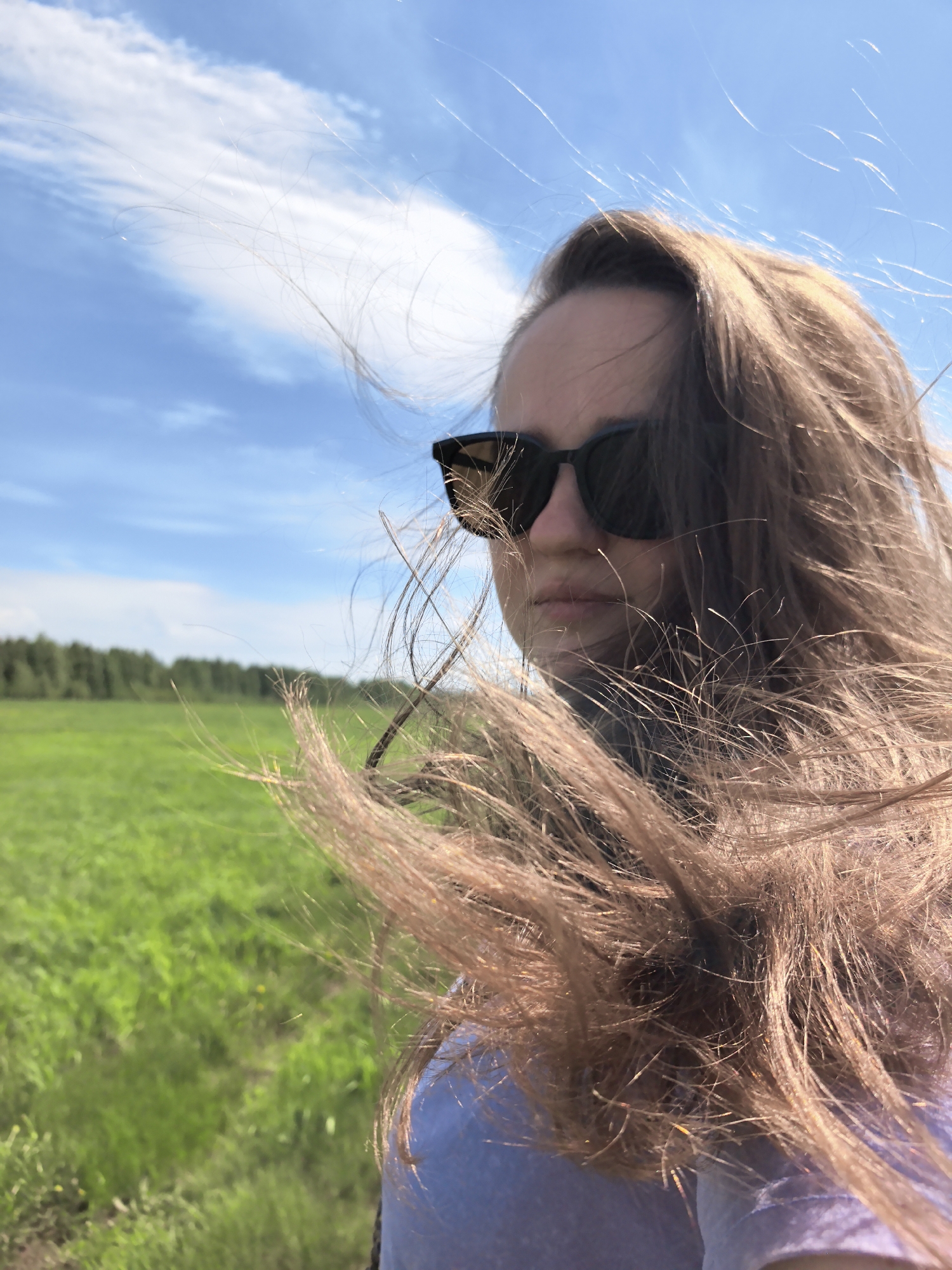 A medium shot depicts a woman with long, light brown hair wearing dark sunglasses, with a field and line of trees visible in the blurred background, taken in Фролы, Russia. Her hair is being blown by the wind, partially obscuring her face. The sky is blue with scattered white clouds. She is wearing a light blue shirt. The field appears to be green with grass.