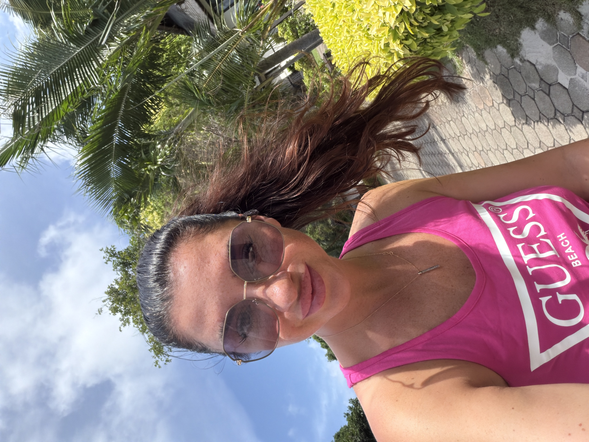 A woman is taking a selfie outdoors in Ao Nang, Thailand, on a sunny day. She is wearing a pink Guess tank top and large, round, rose-tinted sunglasses. Her long, dark brown hair is pulled back into a ponytail and is blowing in the wind. She is wearing a delicate gold necklace with a small, dark pendant. Behind her, a pathway made of interlocking hexagonal stones is visible, bordered by lush green foliage and palm trees. The sky above is bright blue with scattered white clouds. The overall impression is one of a casual, warm-weather outing.