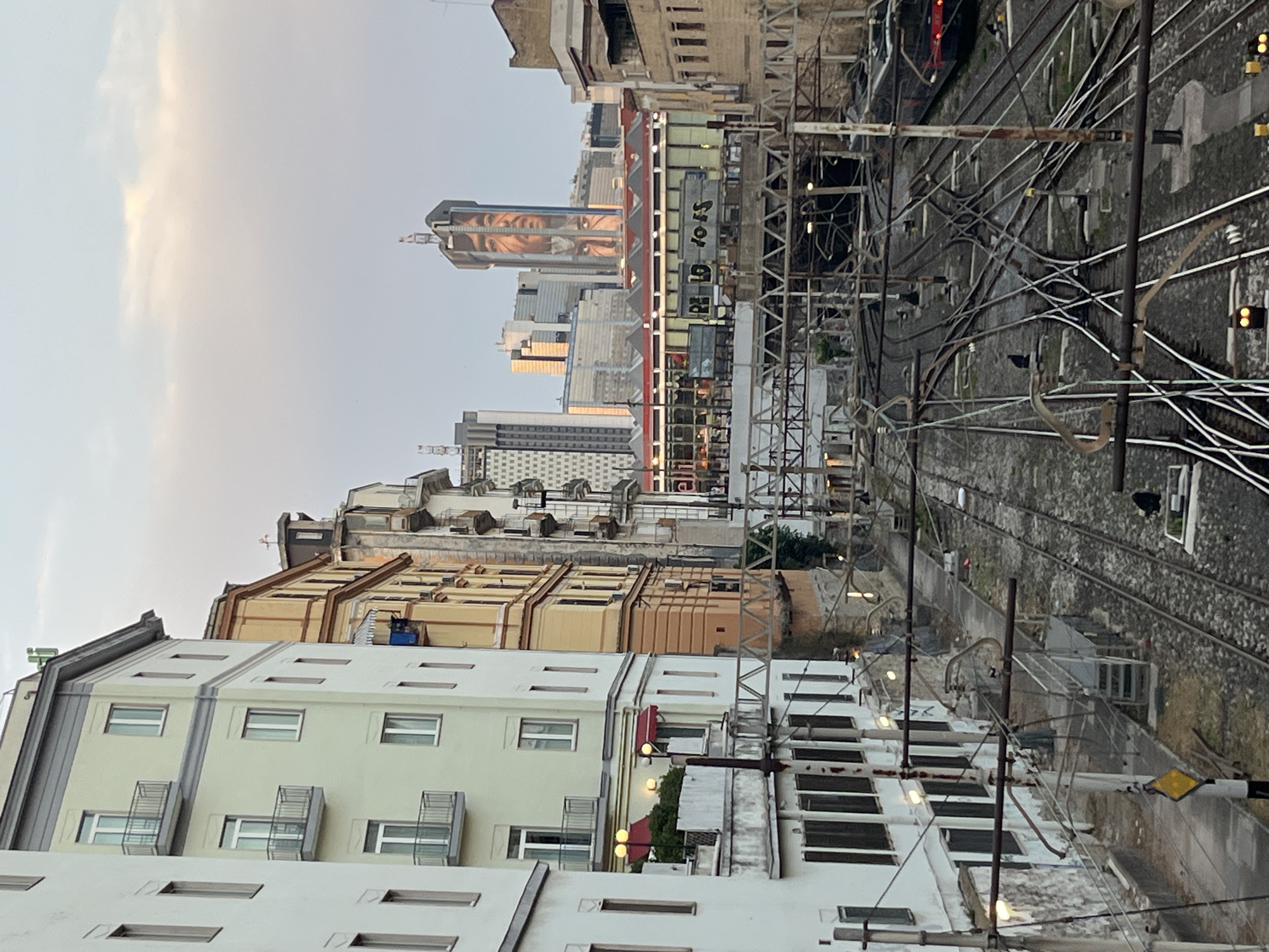 This image depicts a view of Naples, Italy, focusing on a complex railway junction in the foreground and urban architecture in the background. The railway tracks are numerous and intertwined, suggesting a significant transportation hub. Overhead power lines and metal structures associated with the railway system dominate the middle ground.
In the background, a tall skyscraper features a large mural of a face, likely a tribute to Diego Maradona, a legendary football player who lived and played in Naples. Several other modern buildings are visible, interspersed with older, more traditional Italian architecture. The sky above is a soft pastel hue, indicating either dawn or dusk. The scene is devoid of people, emphasizing the infrastructure and the urban landscape. The overall impression is one of a bustling city's functional heart, with artistic expression integrated into its modern skyline.