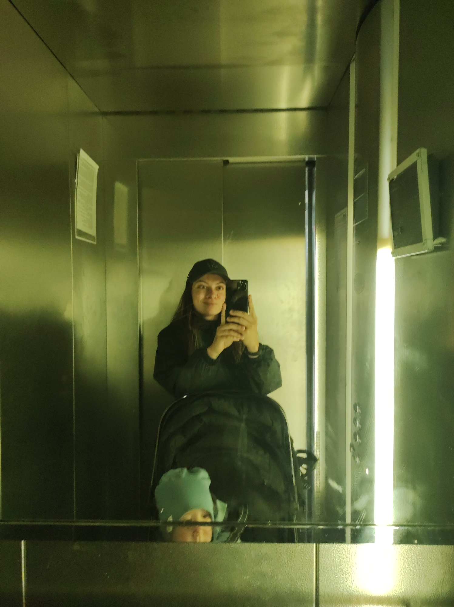 A person in a black jacket and cap is taking a selfie in an elevator. The person is positioned behind a stroller in which a child wearing a light blue hat is seated. The elevator has a stainless steel interior. A fluorescent light fixture is positioned on the right side of the elevator. There is a control panel on the right, with push buttons. There are postings on the left wall of the elevator. The image may have been taken in the city of Chaikovsky, Russia.