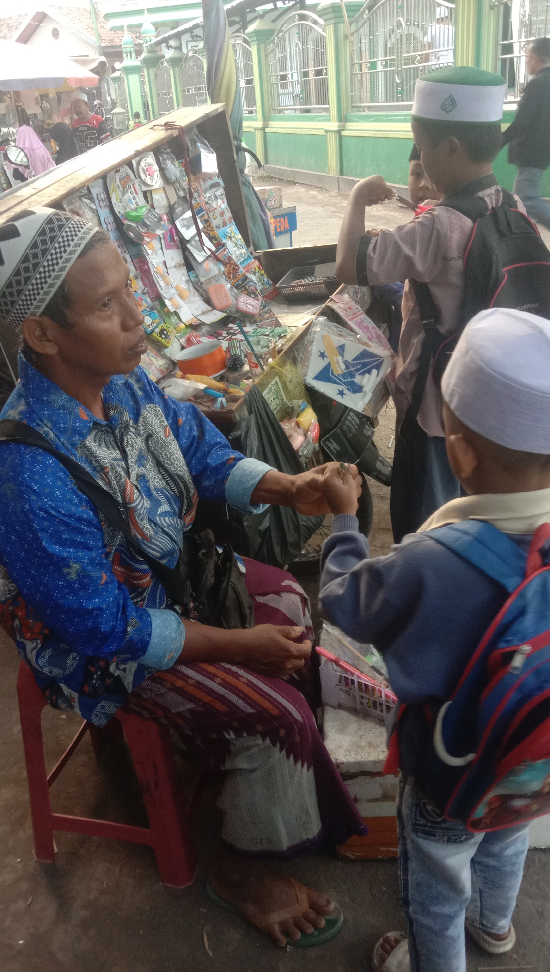 The image shows a street scene in Ngempit, Indonesia, in front of a building with green and white striped walls and a metal fence. A person, possibly a vendor, is seated on a red stool, wearing a blue patterned shirt and a patterned sarong. They are interacting with a young boy wearing a white skullcap, a light brown shirt, and jeans, who has a backpack. Another child, also wearing a skullcap, is partially visible behind the first child, looking towards a stall filled with various items, including toys and snacks. The stall is set up on a wooden cart. The ground is paved with concrete and dirt. The overall atmosphere appears to be a bustling marketplace.