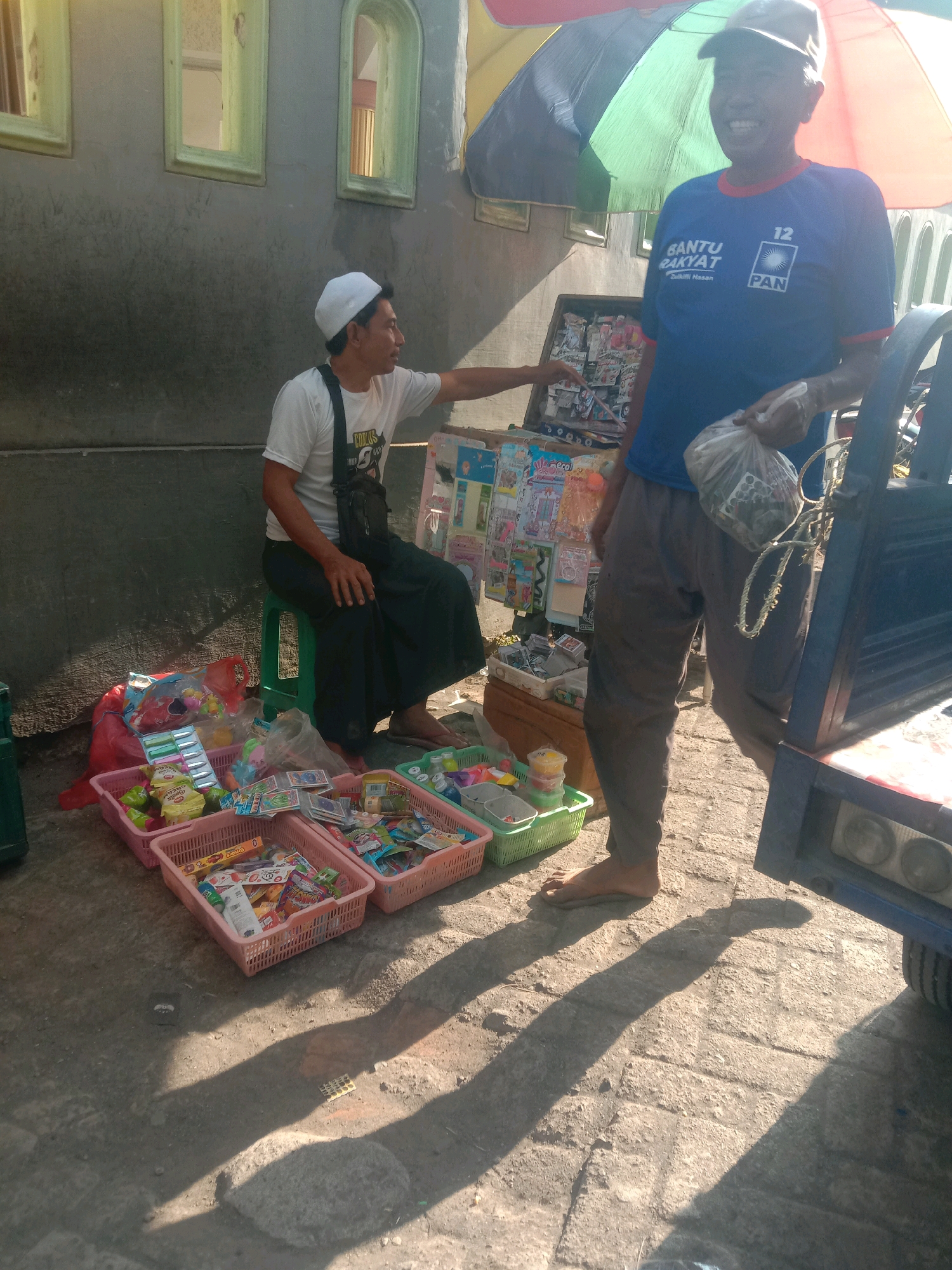 Two men are in an outdoor market stall. One man, wearing a white t-shirt and cap, is seated on a green stool and reaching towards a display of goods. He appears to be interacting with a customer. The second man, wearing a blue t-shirt with Indonesian political party logos and a baseball cap, is standing and holding a plastic bag of items. He is smiling. The stall is filled with various small items, likely toys and snacks, displayed in pink and green plastic baskets. The background shows a grey wall with three windows. Shadows from a large umbrella and the men are cast on the ground. The scene suggests a typical street market setting in Karang Anyar, Indonesia.