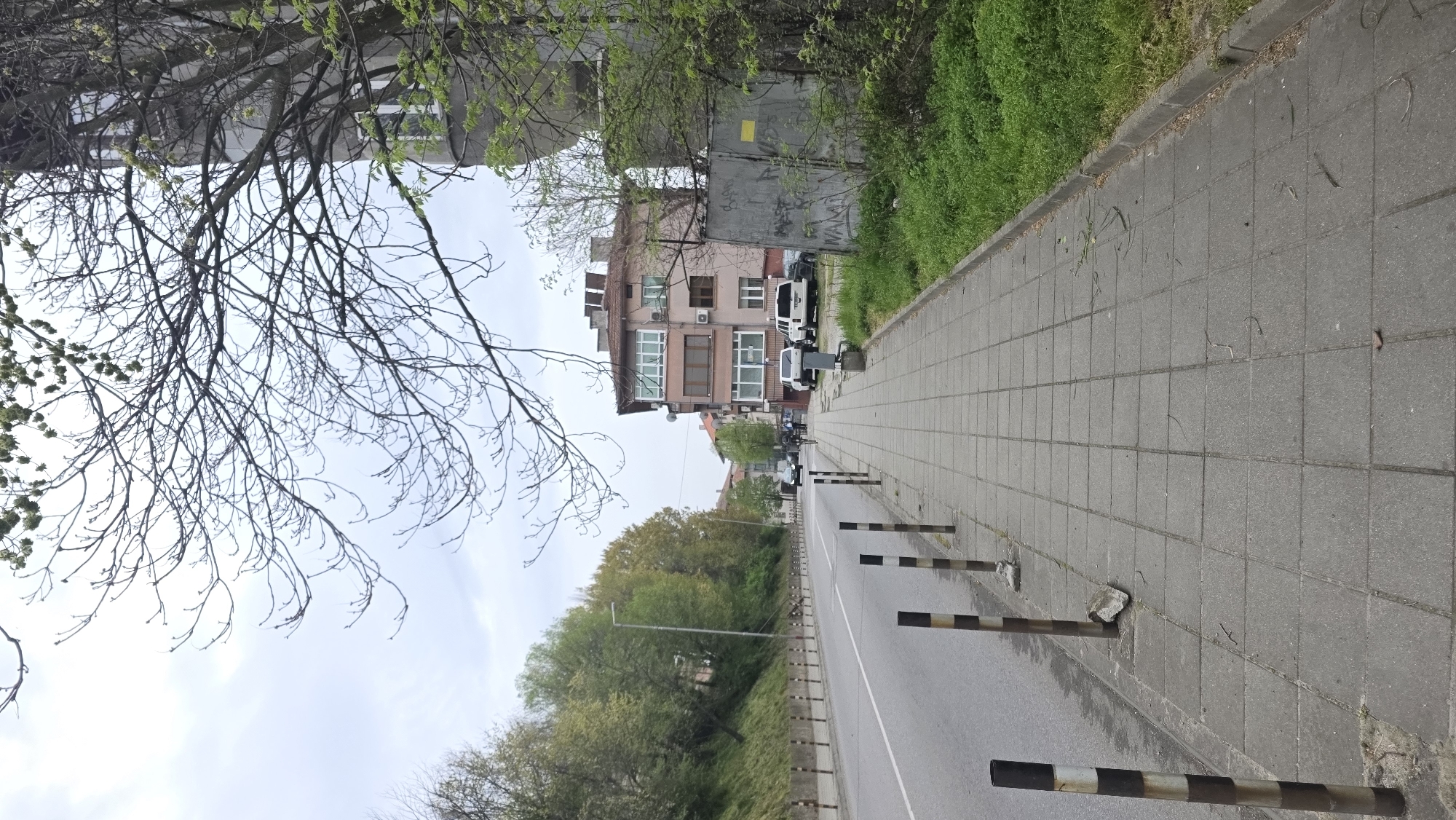 This image depicts a street scene in Blagoevgrad, Bulgaria. A paved sidewalk runs along the right edge of the frame, adjacent to a street. The sidewalk is constructed of square grey paving stones and features a series of striped metal bollards separating it from the road. A building with multiple stories is visible to the right of the sidewalk and road. On the left side of the image, trees with budding leaves obscure part of the sky and a building. The overall impression is of a quiet, urban street in early spring. Several cars are parked along the side of the road.