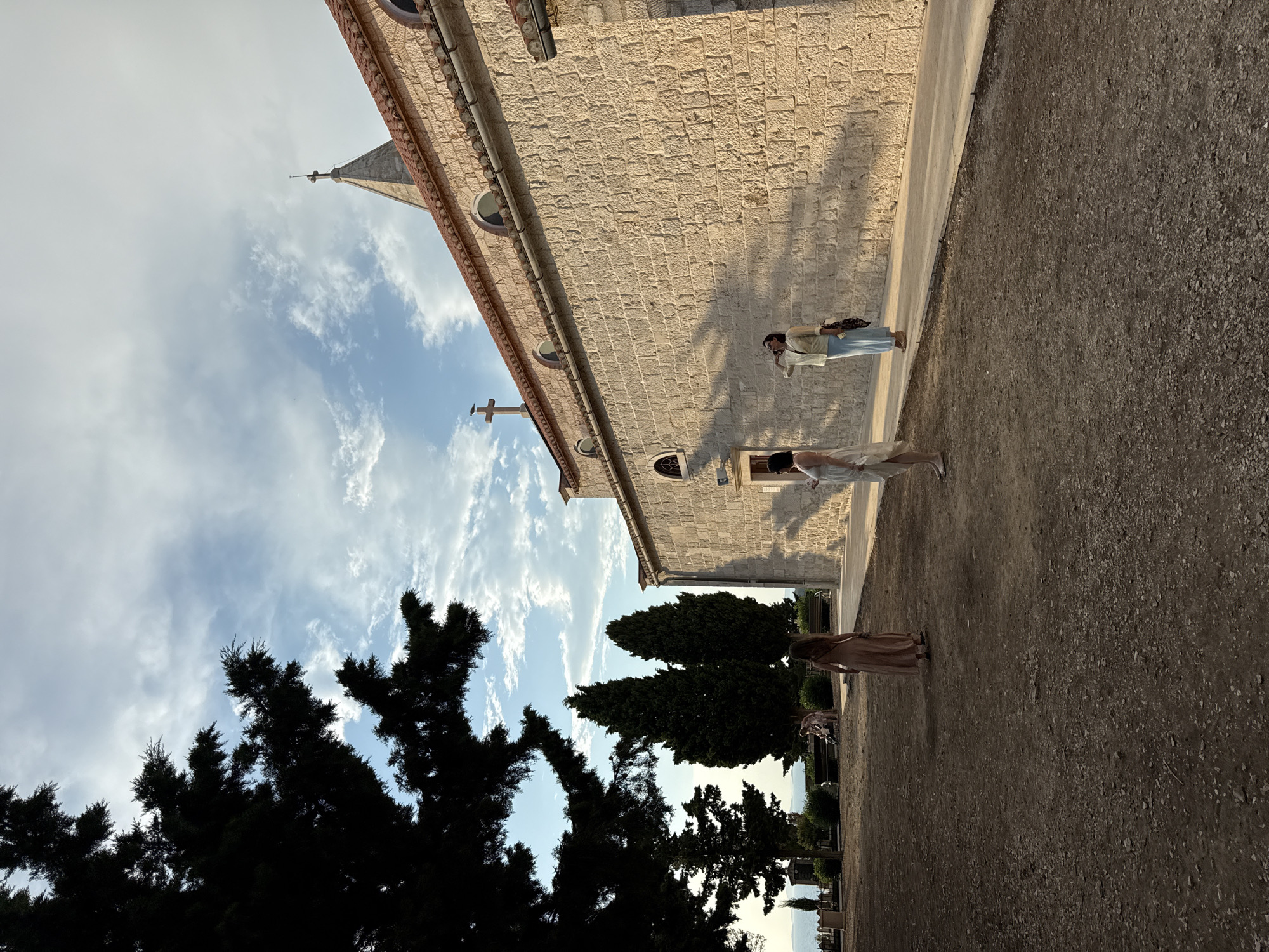 A wide shot captures the exterior of a stone church in Primošten, Croatia, during what appears to be late afternoon or early evening, indicated by the warm sunlight casting long shadows. The church, constructed from light-colored, roughly hewn stones, features a red-tiled roof and a prominent bell tower topped with a cross. Several arched windows are visible along its facade.
In the foreground, a dry, dusty ground is scattered with small stones and pebbles. To the left, the dark silhouette of a large evergreen tree with dense foliage partially obscures the sky. In the middle ground, three women are present. The woman furthest to the left stands with her back to the camera, wearing a long, light-colored dress. Nearer the church, a second woman in a white dress stands facing the church wall, while a third woman, wearing a light yellow blouse and blue pants, stands with her right hand raised to her forehead, shielding her eyes from the sun.
Behind the women, a line of tall, dark green cypress trees forms a backdrop. In the distance, a faint line of the Adriatic Sea and coastline can be seen, suggesting the church is situated on a hill overlooking the water. The sky above is a mix of blue with scattered white and grey clouds. The overall atmosphere is serene and picturesque, capturing a moment of quiet contemplation or sightseeing.