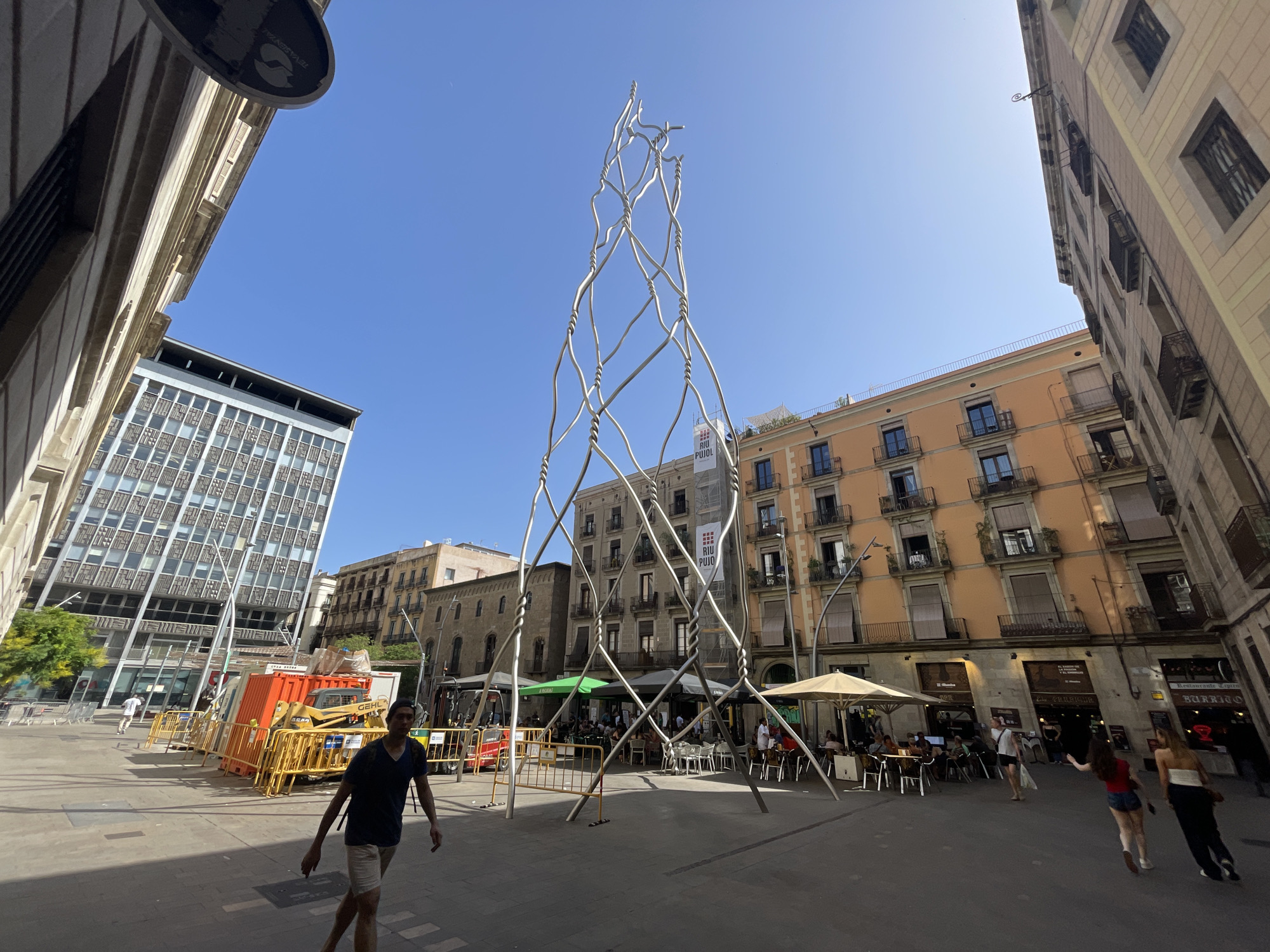 The image captures a vibrant outdoor scene in Barcelona, Spain, bathed in bright sunlight, suggesting it's daytime on a clear, sunny day. The central focus is a tall, abstract metal sculpture that resembles a twisted, woven tower. Surrounding the sculpture is a public square with several buildings.
On the left, a modern, grey building with a grid-like facade stands out. To the right and in the background, older buildings with balconies and numerous windows are visible, some painted in warm, earthy tones. Some of these buildings house businesses, with visible signage.
In the foreground, a man wearing a dark t-shirt and light-colored shorts walks towards the camera. Other people are scattered throughout the square, some seated at outdoor cafes under umbrellas and awnings, enjoying refreshments. Construction elements, including an orange container and yellow barriers, are present in the middle ground, indicating potential ongoing work.
The overall atmosphere appears relaxed and lively, with people engaging in everyday activities in an urban setting. The architecture and the presence of outdoor dining suggest a typical European city square. The clear blue sky and strong sunlight contribute to a sense of warmth and activity. A banner with "RIU PUIG" is visible on one of the buildings.