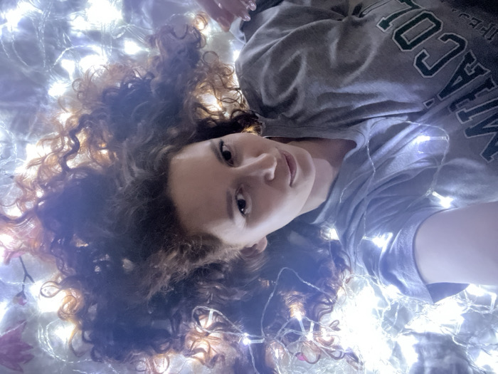 A female individual with voluminous, curly, reddish-brown hair is depicted in a close-up, high-angle shot, lying on her back. She wears a grey t-shirt with visible green lettering, partially spelling "COLLEGE". Her gaze is directed towards the camera. She is positioned amidst a dense arrangement of numerous small, bright white LED string lights, which are intertwined with her hair and draped across her upper body. Faint elements resembling reddish-orange autumn leaves are interspersed within the lights, particularly in the upper left quadrant of the frame. The immediate environment is illuminated primarily by these diffuse LED lights. The specific location within Saint Petersburg, Russia, cannot be determined from the visual information provided.