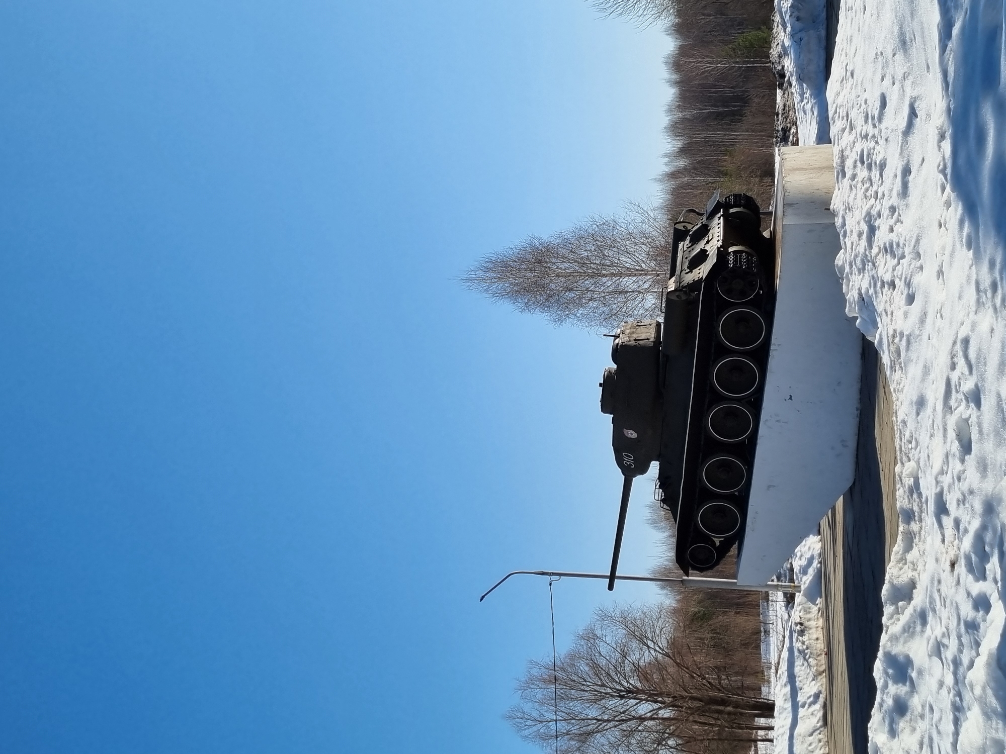 An outdoor photograph shows a Soviet tank displayed on a concrete platform on a winter day. The tank, dark green, is positioned parallel to the platform's edge, and has "310" painted in white on its side. The cannon extends towards the clear blue sky. The platform is covered with snow. Beyond the platform is a partially snow-covered area with bare trees in the background. A tall metal pole stands next to the tank. The photograph was taken in Марковский, Russia.