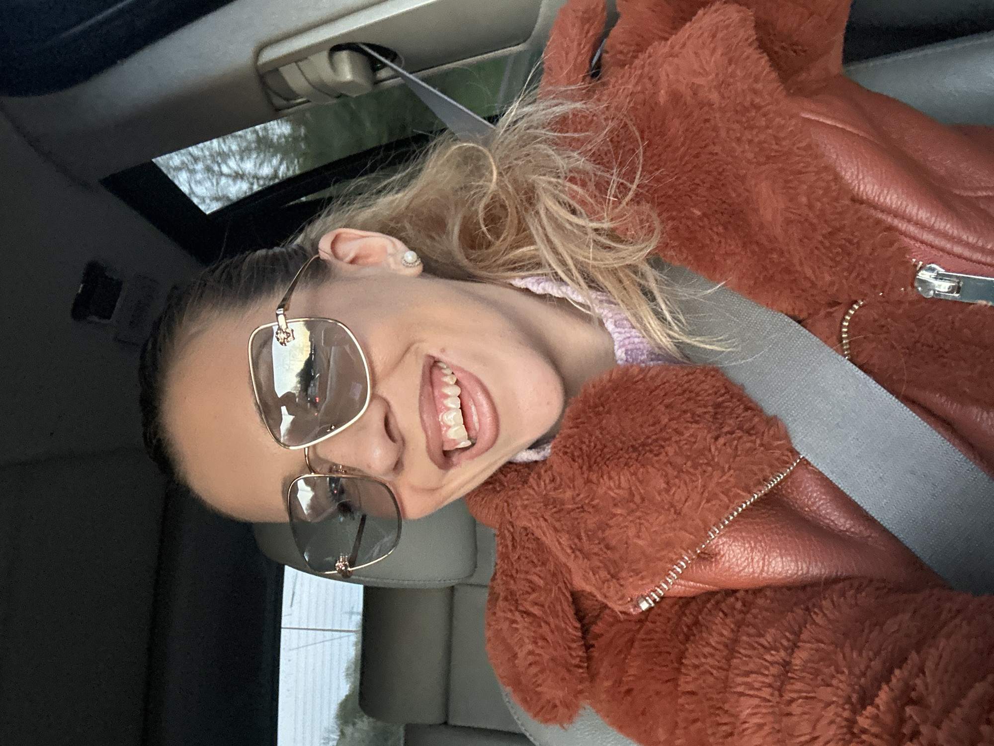 A woman is in a car, facing the camera at an angle, wearing a gray seatbelt, sunglasses, and a reddish-brown jacket with both furry and leather-like textures. She is smiling and exposing her teeth. There is a sunroof above. The location cannot be determined to be Grays, United Kingdom from the provided image.