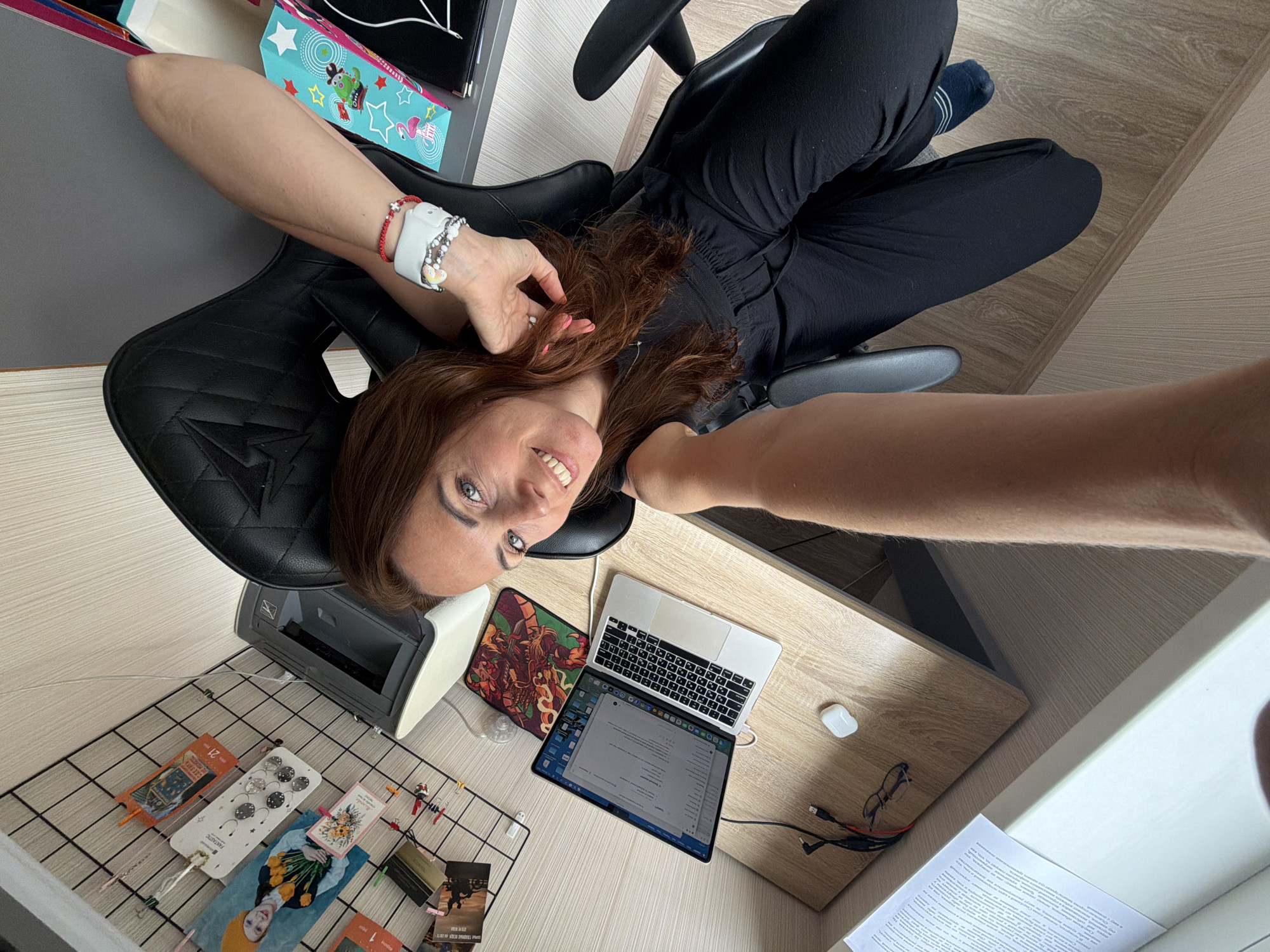 The image depicts a woman sitting at a desk in what appears to be a home office or workspace. She is looking at the camera and smiling, with her head tilted back and arms resting on the chair. On the desk, there is an open laptop, a mouse, a pair of glasses, and charging cables. To the left of the desk, a black wire grid is adorned with various items including photos, bookmarks, and clips. In the background, a printer and a colorful gift bag are visible. The woman is wearing black clothing and blue socks. The environment suggests a casual work setting. There are no specific visual cues within the image that directly indicate a location in Birobidzhan, Russia.