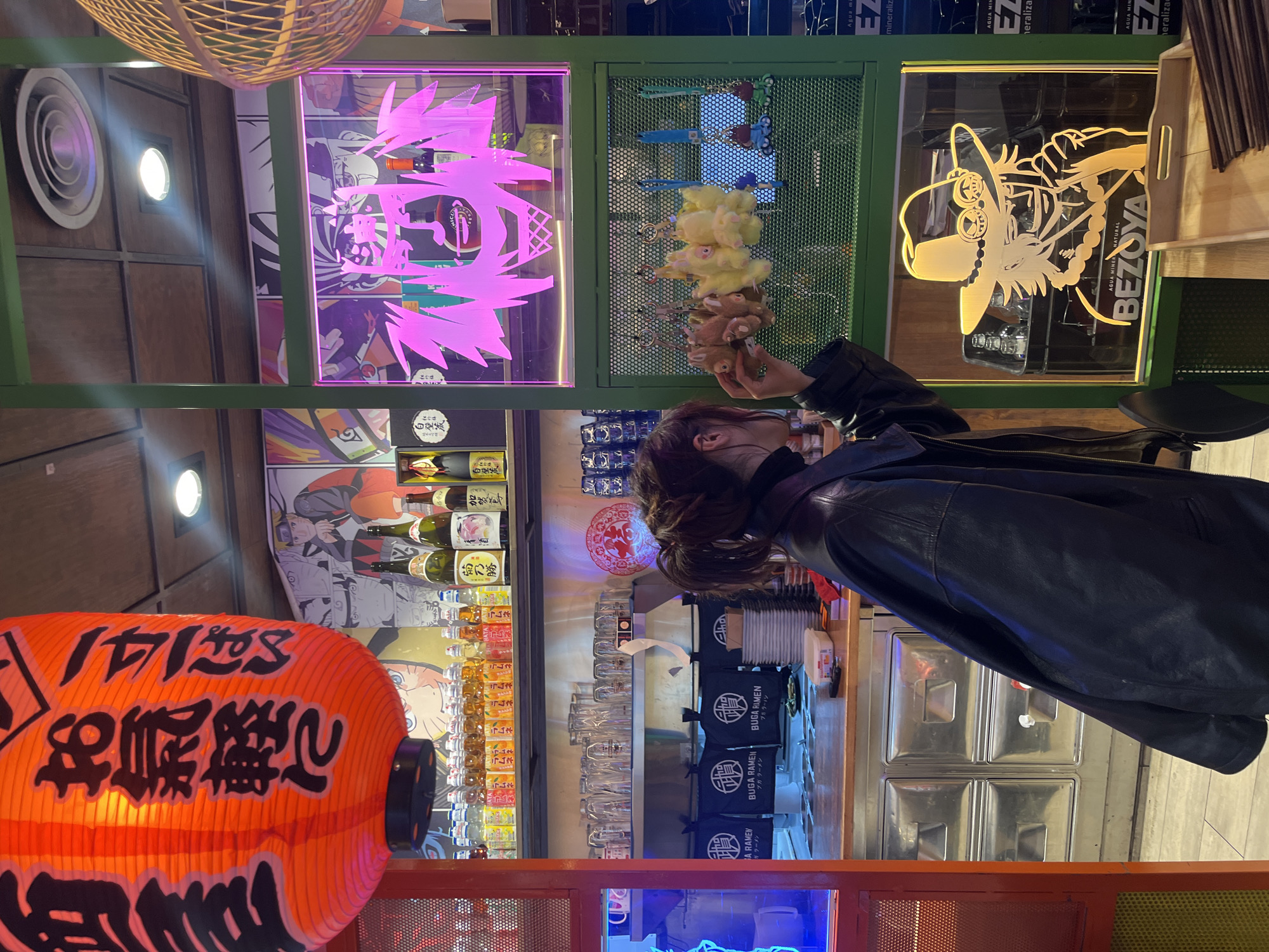 The image depicts the interior of a restaurant, potentially located in Barcelona, Spain. A woman is visible in the foreground, near a display of stuffed animal keychains, specifically teddy bears, arranged on a green metal grid panel.
A lit orange lantern, with traditional Japanese writing on it, hangs prominently on the left. Behind the counter, there are rows of glasses, small bottles of drinks, and storage containers, along with menus. The restaurant walls are decorated with various posters, including anime/manga-style art with neon pink outlines, and brand logos.