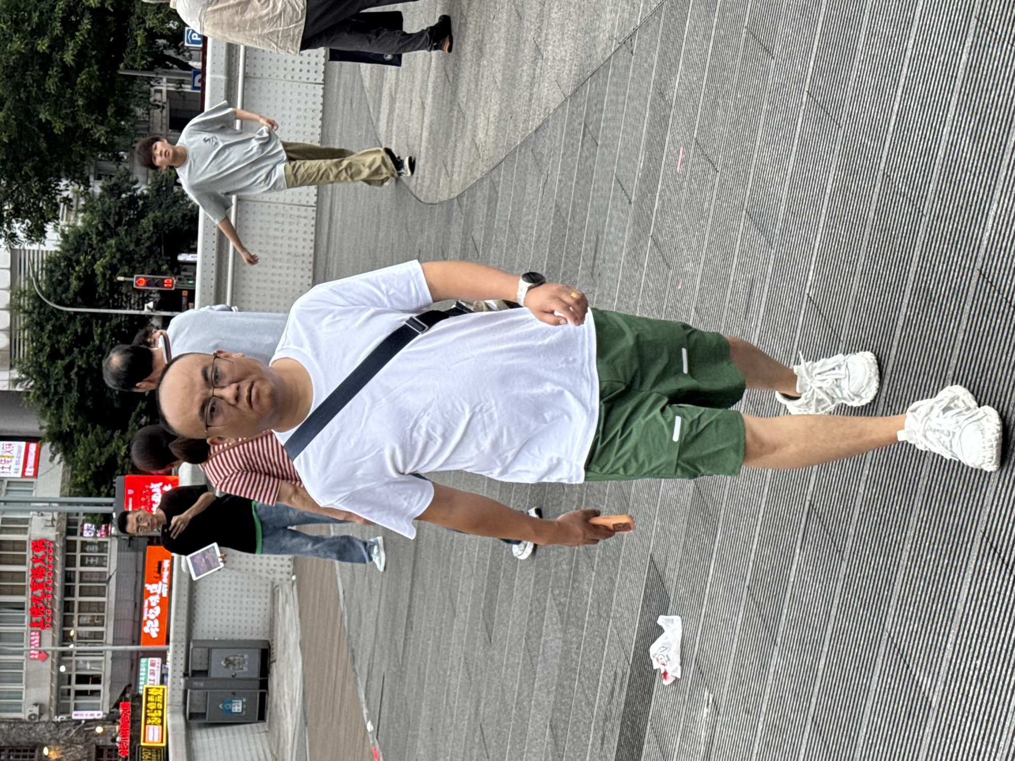 A medium shot captures a bald, middle-aged East Asian man with glasses walking forward on a slatted pedestrian walkway. He is wearing a white t-shirt, green shorts, and white sneakers, with a black strap bag slung across his chest and a smartwatch on his left wrist. His right hand is extended, holding an orange object that appears to be a phone. To his left, a white plastic bag lies on the walkway. In the background, several other pedestrians are visible, including a young East Asian man in a grey t-shirt and khaki pants walking away from the camera. The environment appears to be an outdoor urban setting, possibly in Yuzhong District, China, with buildings and trees in the distance.