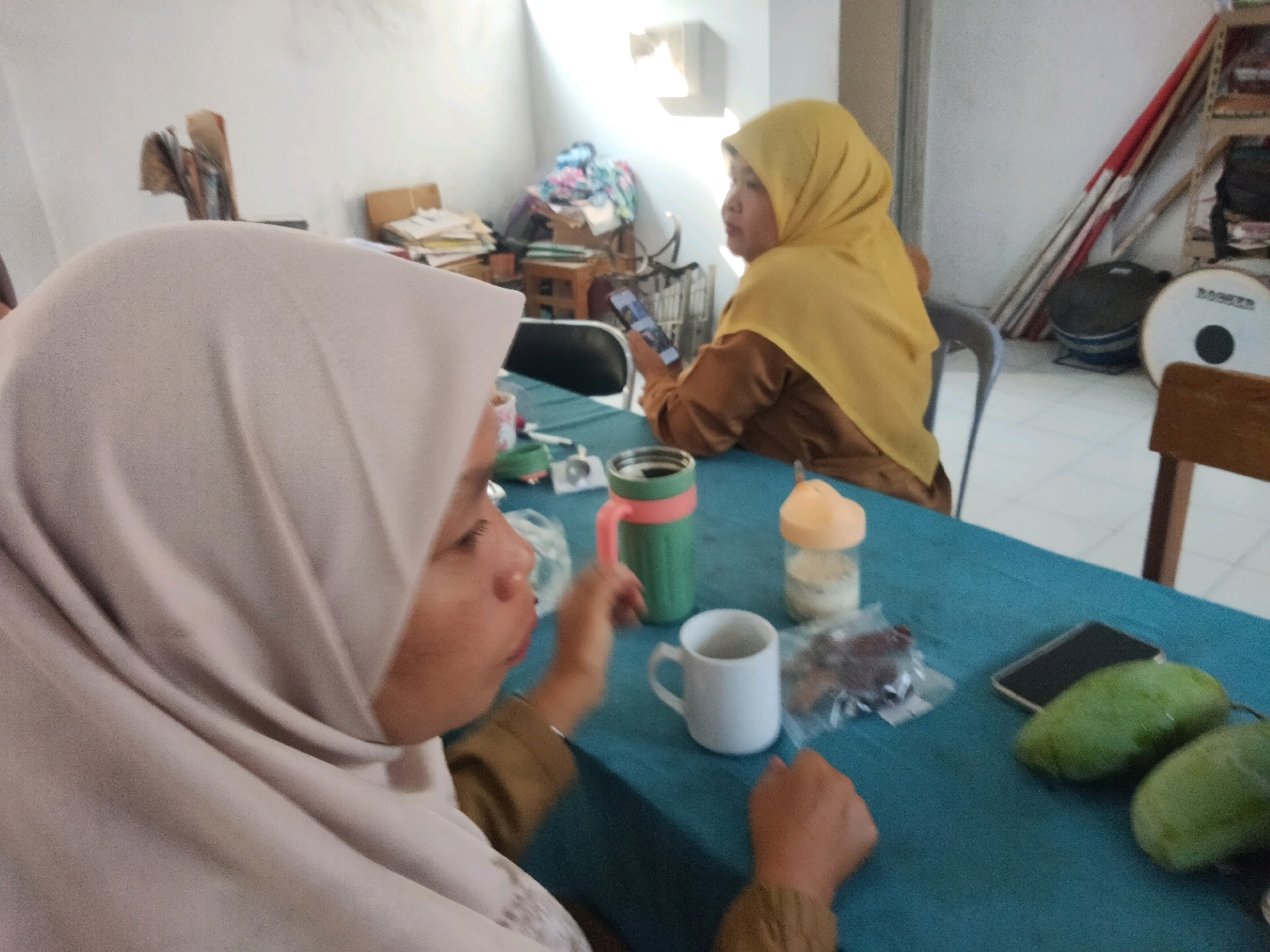 In Suka Sari, Indonesia, two women are seated at a blue-clothed table. The woman in the foreground wears a light-colored hijab and is partially visible. The woman in the background wears a yellow hijab and a brown top. She is holding a smartphone. On the table are various items: a green thermal mug with a pink lid, a white mug, a clear bottle with an orange lid and white contents, a plastic-wrapped package of food, a smartphone, and two green mangoes. In the background, shelves hold items and two large cylindrical objects, one white with black markings. The environment appears to be an indoor space with white walls and a tiled floor.