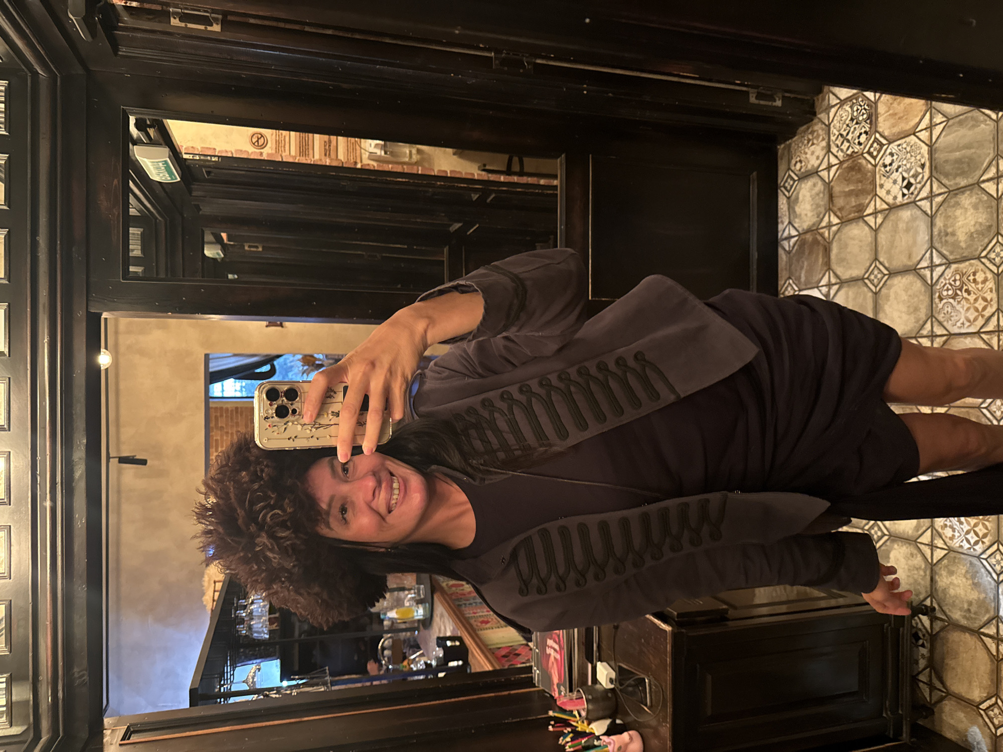 The image shows a woman taking a selfie in a dimly lit hallway with mirrored doors. She is wearing a dark dress and a dark jacket with ornate braiding. She is also wearing a large, dark, fluffy hat. The hallway has patterned tile flooring and dark wood paneling. In the reflection of the mirrored doors, there are glimpses of what appears to be a bar or restaurant area, with shelves of glasses and bottles visible. There are also signs in Russian visible in the reflection. The overall impression is of an interior space, possibly a private club or restaurant, in a city known for its ornate architecture. Given the Russian signage, the location is likely Saint Petersburg, Russia.