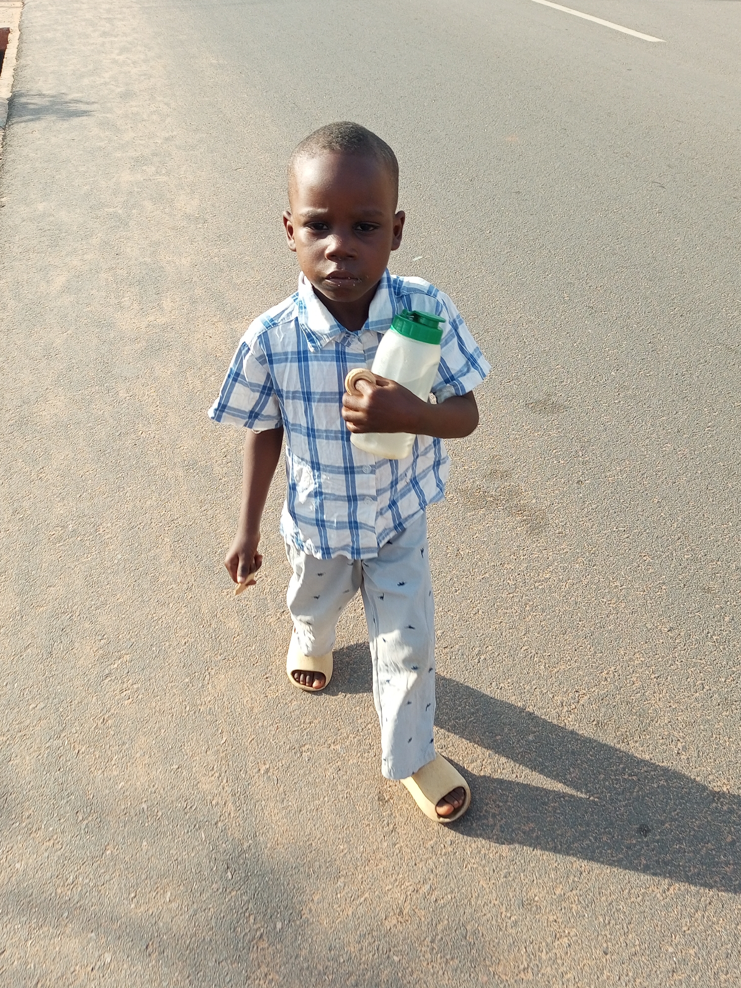 A young Black boy, likely under ten years old, walks on a paved road in Tempure, Nigeria. He is wearing a blue and white checkered short-sleeved shirt and light-colored pants with small dark blue patterns. On his feet are light brown slip-on sandals. The boy is holding a white water bottle with a green cap in his right hand. The road is asphalt, with a lighter colored shoulder on the left side. A white line is visible on the road in the background. The scene is bathed in sunlight, casting a long shadow of the boy to his right.