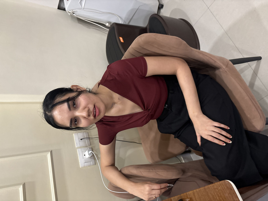 The image displays an indoor scene within a commercial establishment, likely a salon or spa, located in Howrah, India. A single adult female is seated towards the center-left in a light beige upholstered chair featuring dark legs. She wears a short-sleeved, ribbed, V-neck maroon top with a twisted front detail and black wide-leg trousers. Her dark hair is styled away from her face, and she possesses light-colored nail polish on her fingernails, with a ring on the fourth digit of her left hand. Her right arm rests on the chair's back, and her left hand is positioned on her lap. She is looking directly at the camera with a neutral gaze.

The background wall is light beige with a rectangular decorative molding present on the upper left side. Two white electrical wall outlets are visible on the wall behind her, with a white charging block plugged into the left outlet and a white cable extending downwards. A portion of a wooden-surfaced table is visible on the far left. To the right of the seated individual, a dark brown, two-tiered unit with a black top surface, resembling a foot spa and featuring an orange "merop" label, is present. Further to the right, a tall, white piece of equipment with white cables is partially visible. The floor consists of light-colored tiles.