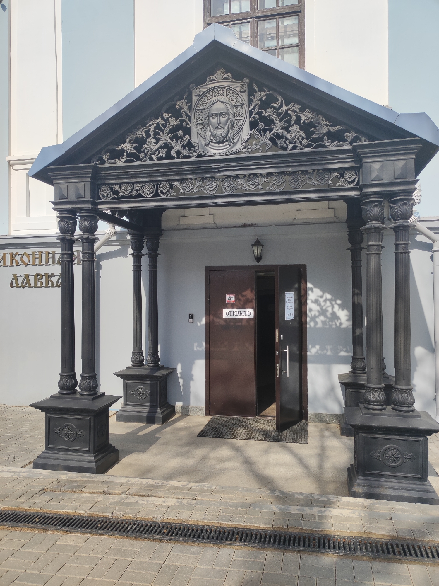 The image captures the entrance to a building, possibly a shop or a religious institution, located in Votkinsk, Russia. The architecture is characterized by a covered portico featuring decorative black metal columns with ornate bases and capitals. Above the doorway, a triangular pediment displays a relief carving of a face, likely depicting a religious figure, surrounded by floral motifs.
The door is ajar, revealing a dark interior. A sign reading "ОТКРЫТО" (OPEN) is affixed to the closed portion of the door. The exterior wall is painted in muted tones of white and light blue, and gold lettering is visible on the left, presumably indicating the name of the establishment.
The ground is paved with gray stone tiles, and a drainage grate runs along the bottom of the frame. The overall scene is bathed in natural light, casting shadows on the facade and the ground. No people are visible in the image.