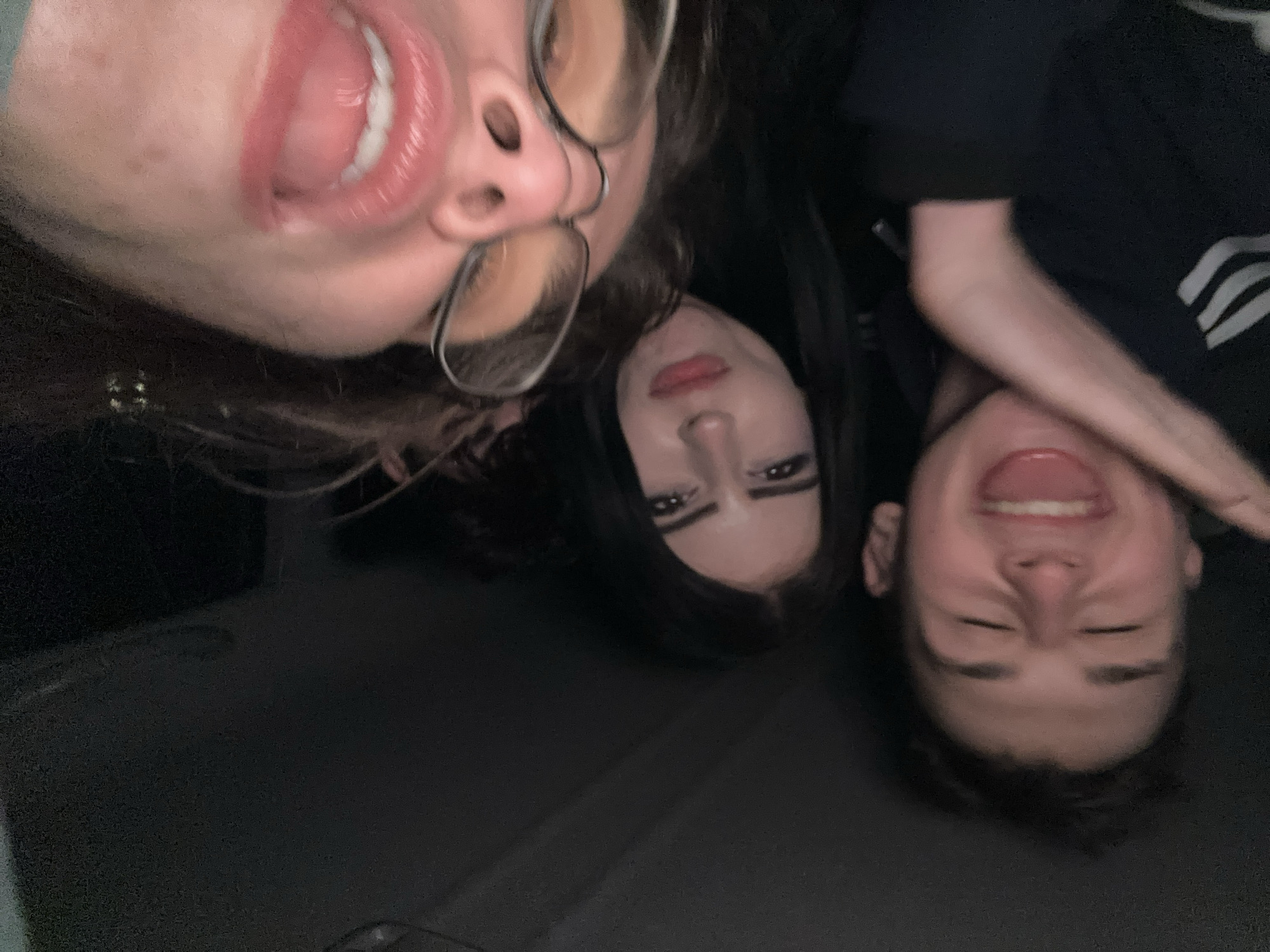 The image shows three individuals. At the top left, a person is visible with an open mouth, glasses, and brown hair. In the center, there is a person with dark hair and makeup. To the right, a person is smiling, wearing a black top with white stripes on the sleeve. The setting appears to be indoors, with a dark-colored background. There is no information to suggest this image was taken in Pleven, Bulgaria.