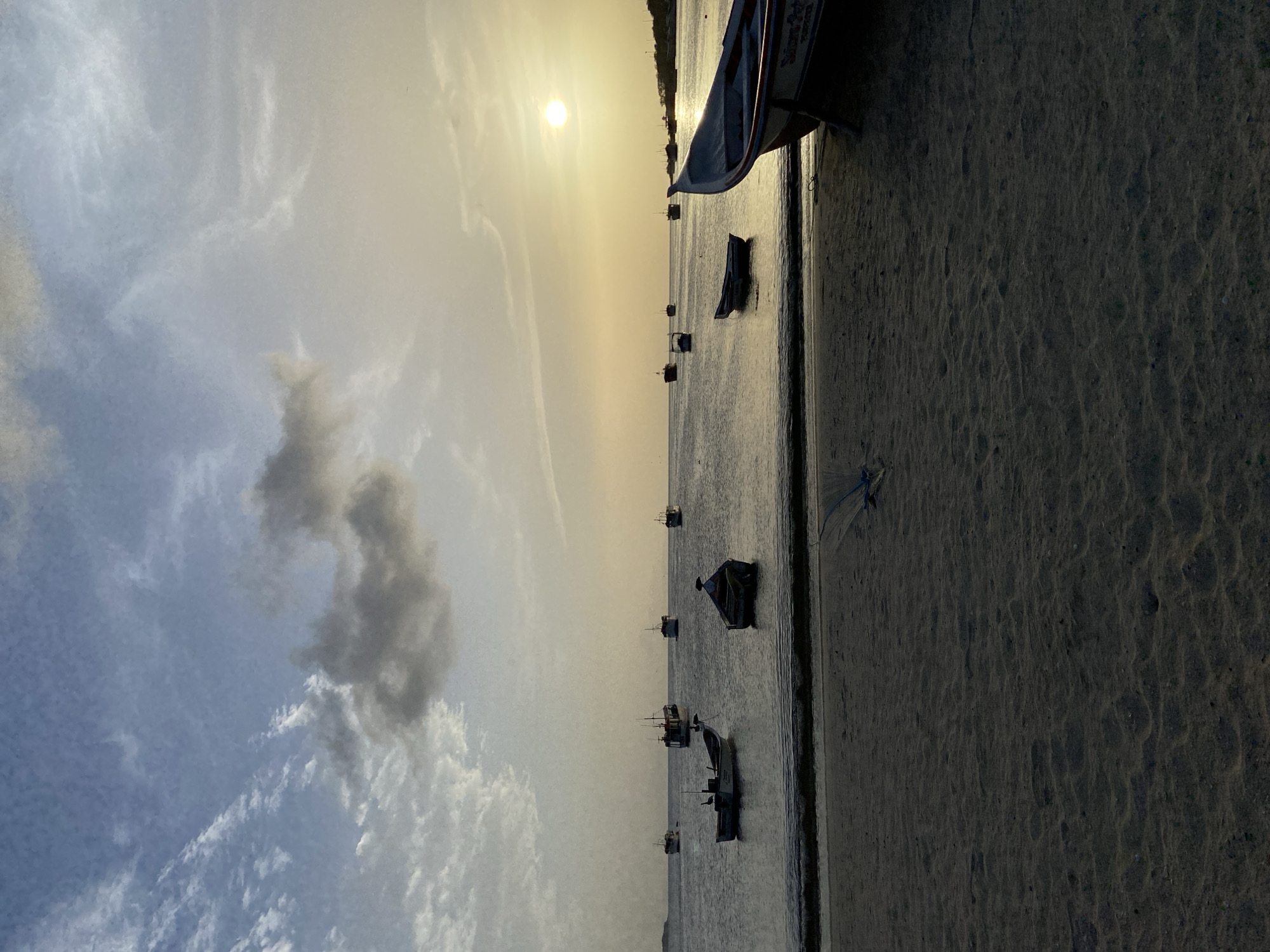 This is a medium shot, taken at eye level, of a beach in Juan Griego, Venezuela, at sunset. The sky is a gradient of blues and yellows, with scattered clouds. The sun is low on the horizon, casting a warm glow over the scene. In the foreground, a sandy beach stretches across the bottom of the frame, with some seaweed and debris visible. A few boats are pulled up onto the sand, including a large fishing boat on the right side of the image. In the middle ground, several smaller boats are moored in the shallow water, some with people in them. In the background, more boats are visible on the horizon, and a distant landmass can be seen. The overall impression is one of peace and tranquility, with the gentle lapping of waves and the warm glow of the setting sun.