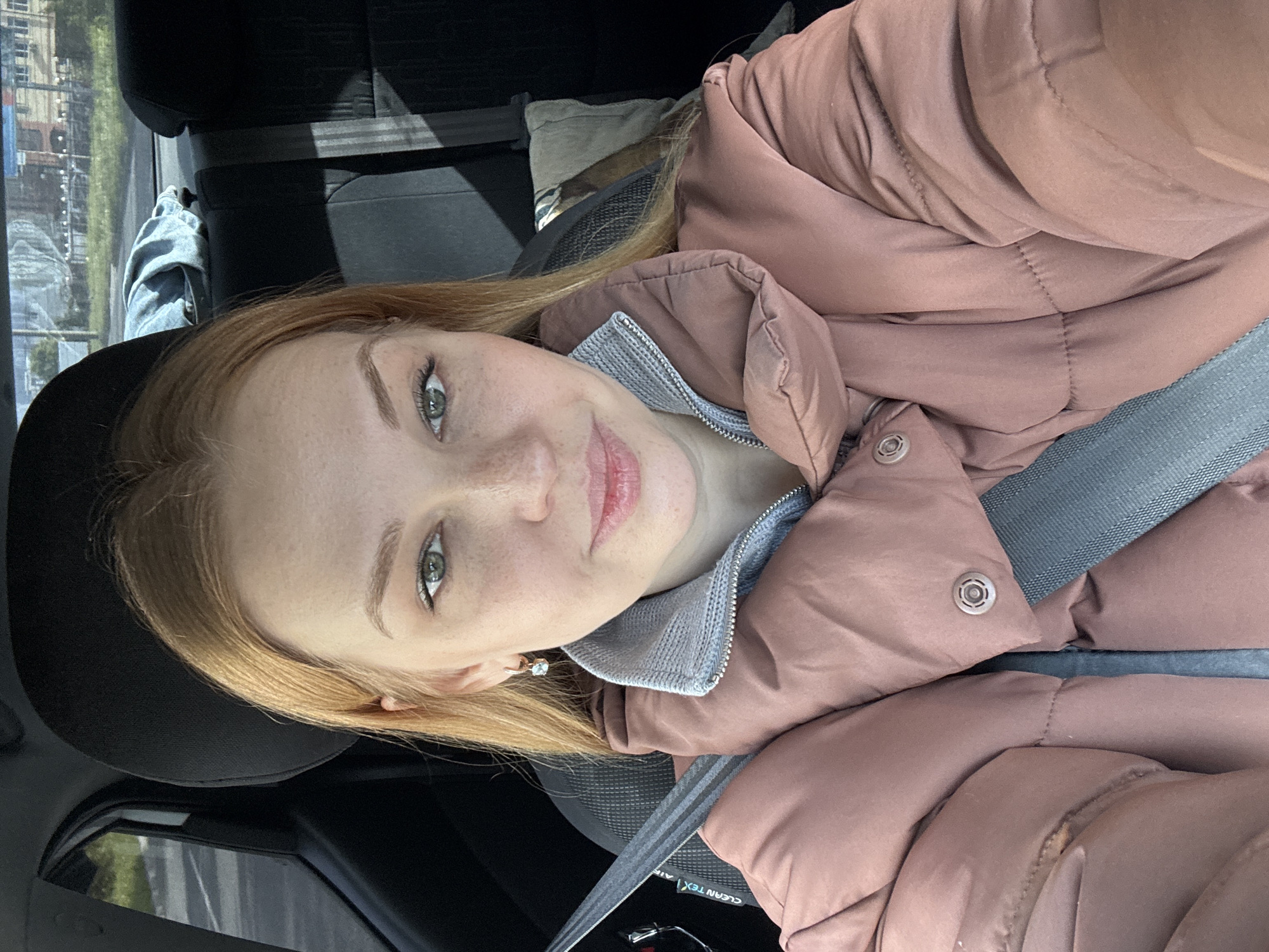 A young woman with light red hair and freckles is in the driver's seat of a car. She is wearing a light pink puffer jacket and a grey top. A grey seatbelt is fastened across her chest. She is looking at the camera with a slight smile. The car is parked, and a view of a building and street can be seen through the side window. The location is unknown.