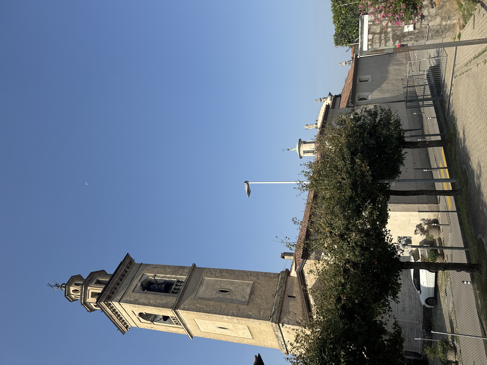 In the city of Balerna, Switzerland, a bell tower stands tall against a clear blue sky. The tower, made of aged stone, features arched openings where bells are visible and a decorative crown at its apex. A crescent moon is visible in the sky to the right of the tower. In front of the tower, a church building with a tiled roof is partially obscured by trees. A white car is parked in front of the church, behind a chain-link fence. The ground is paved with light-colored stones, and a sidewalk runs along the fence. To the right of the church, a wall covered in ivy and blooming pink flowers adds a touch of color to the scene. A street lamp stands near the church, its light directed downwards. The overall impression is of a peaceful, sunny day in a historic town.